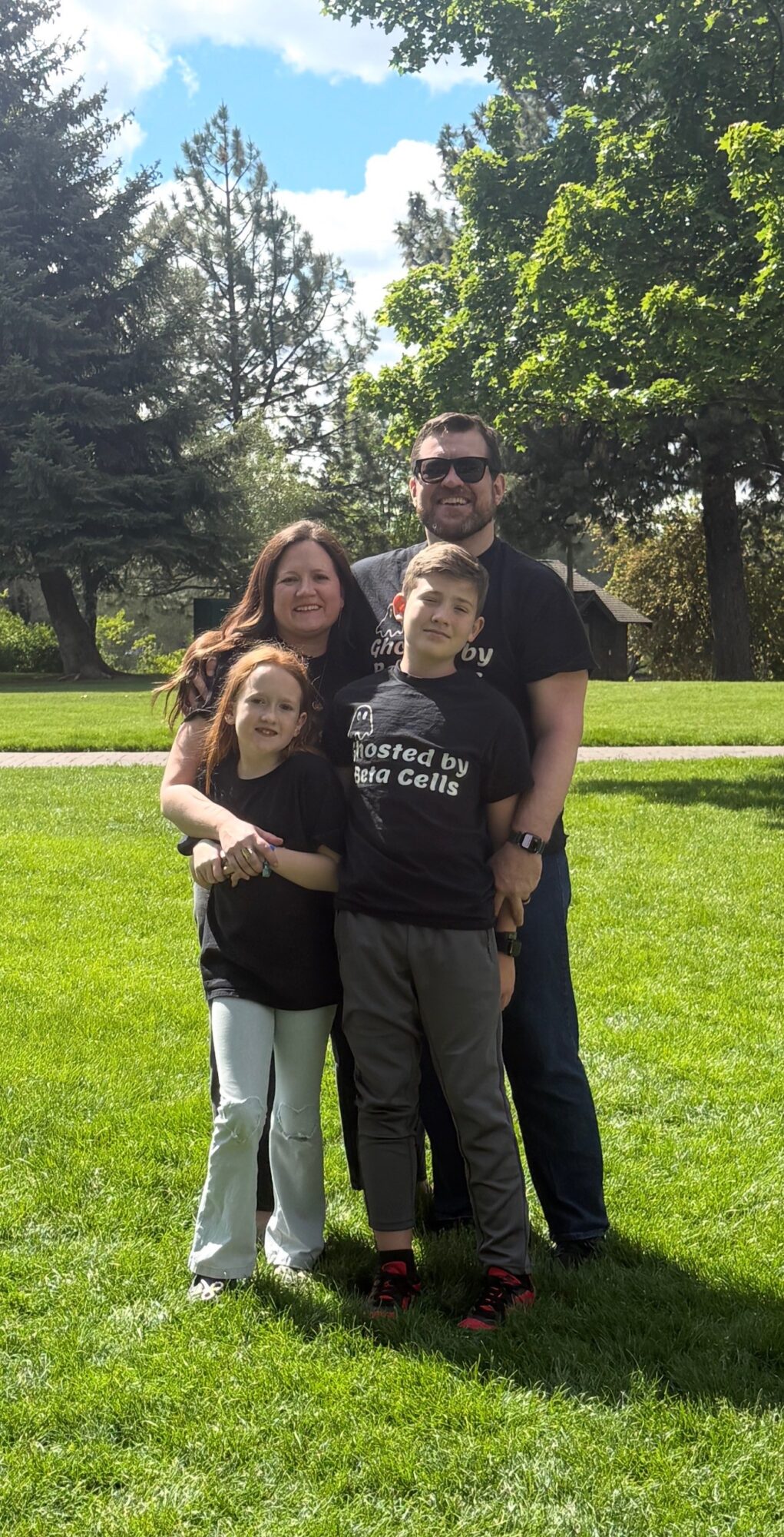 Family of four standing outdoors on grass with trees and blue sky, smiling at the camera.