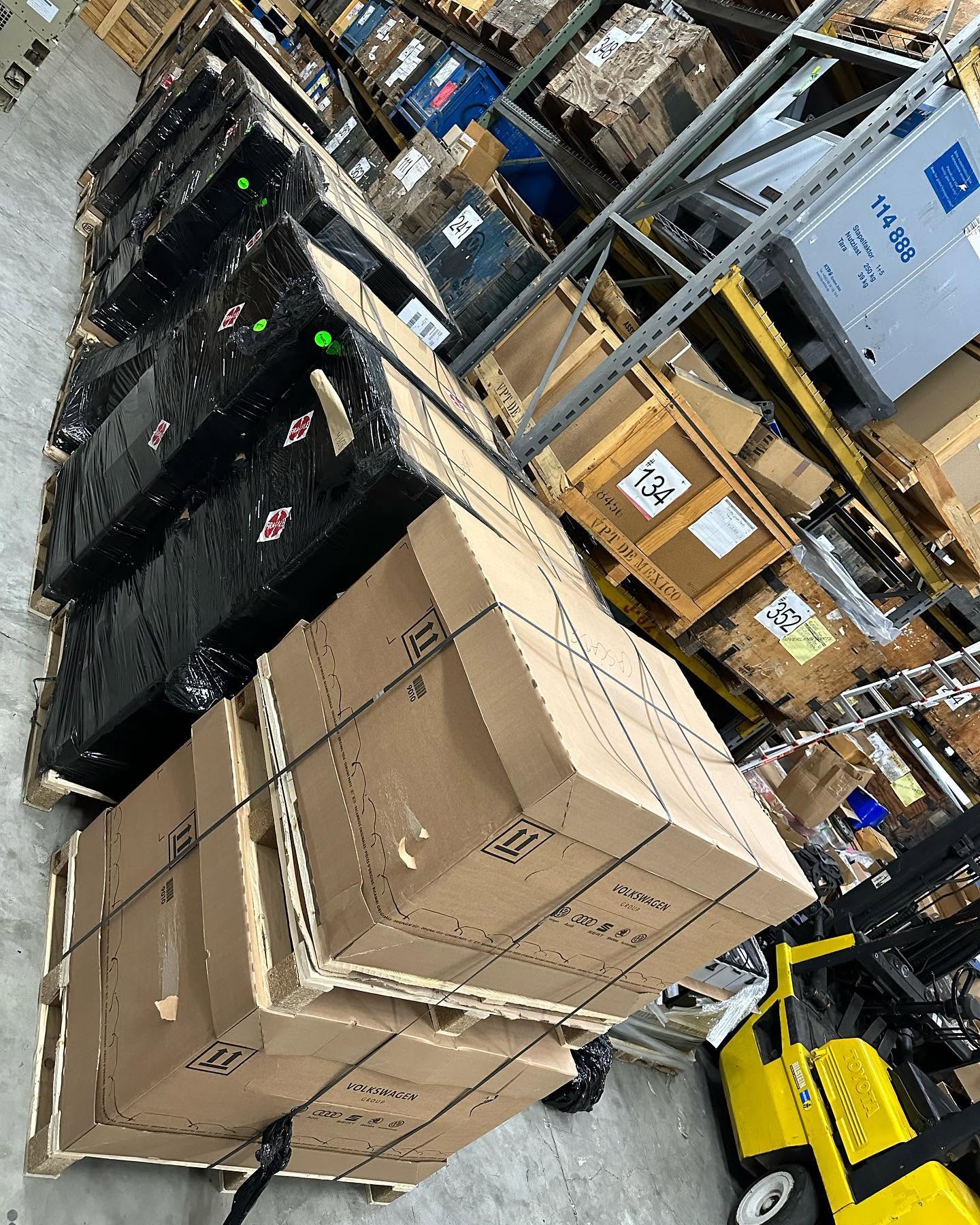 Stacked large cardboard boxes and black electronic devices on pallets in a warehouse.