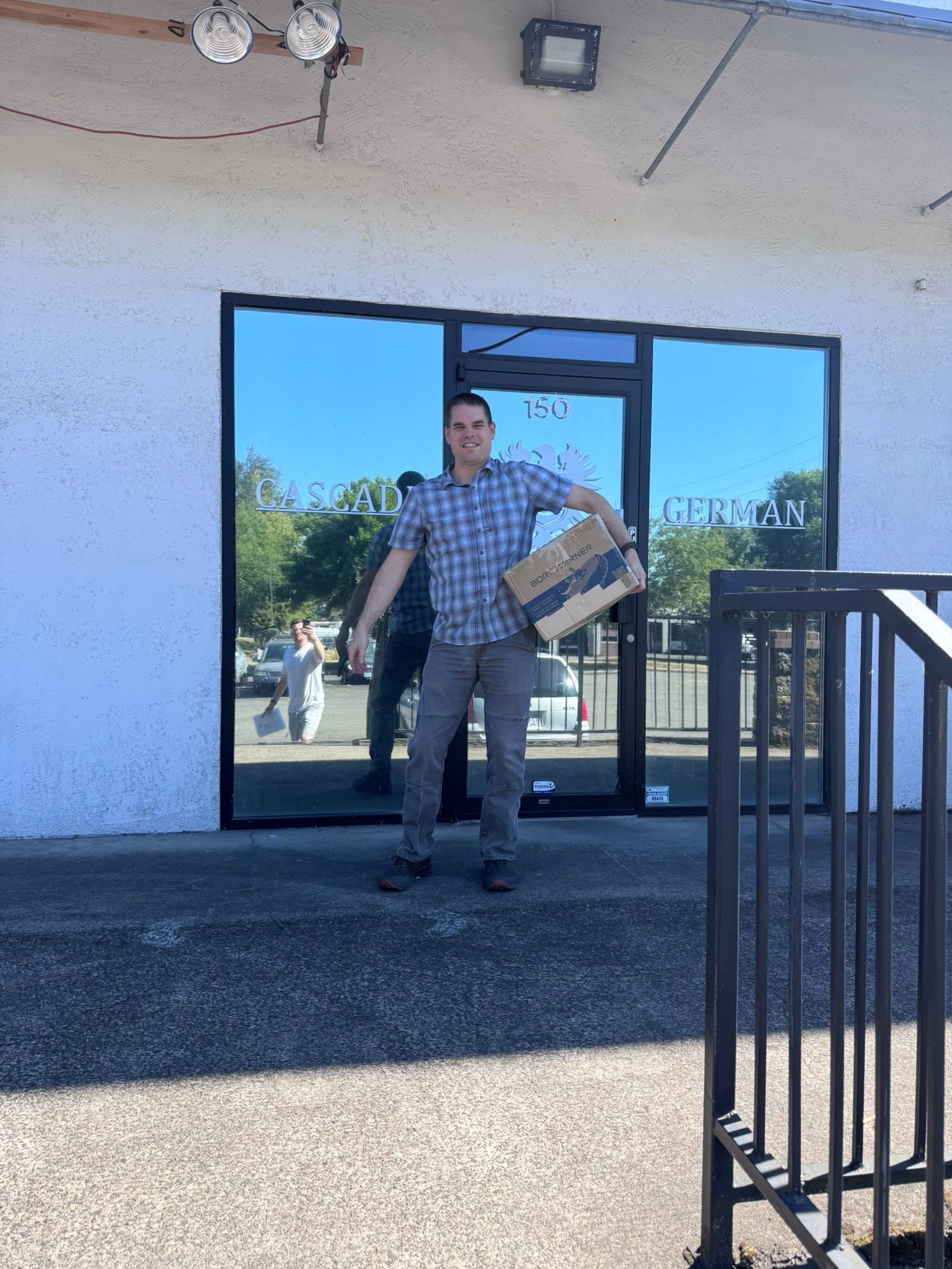 Man standing outside a glass door holding a box, reflected in the glass, with trees and a building in the background.