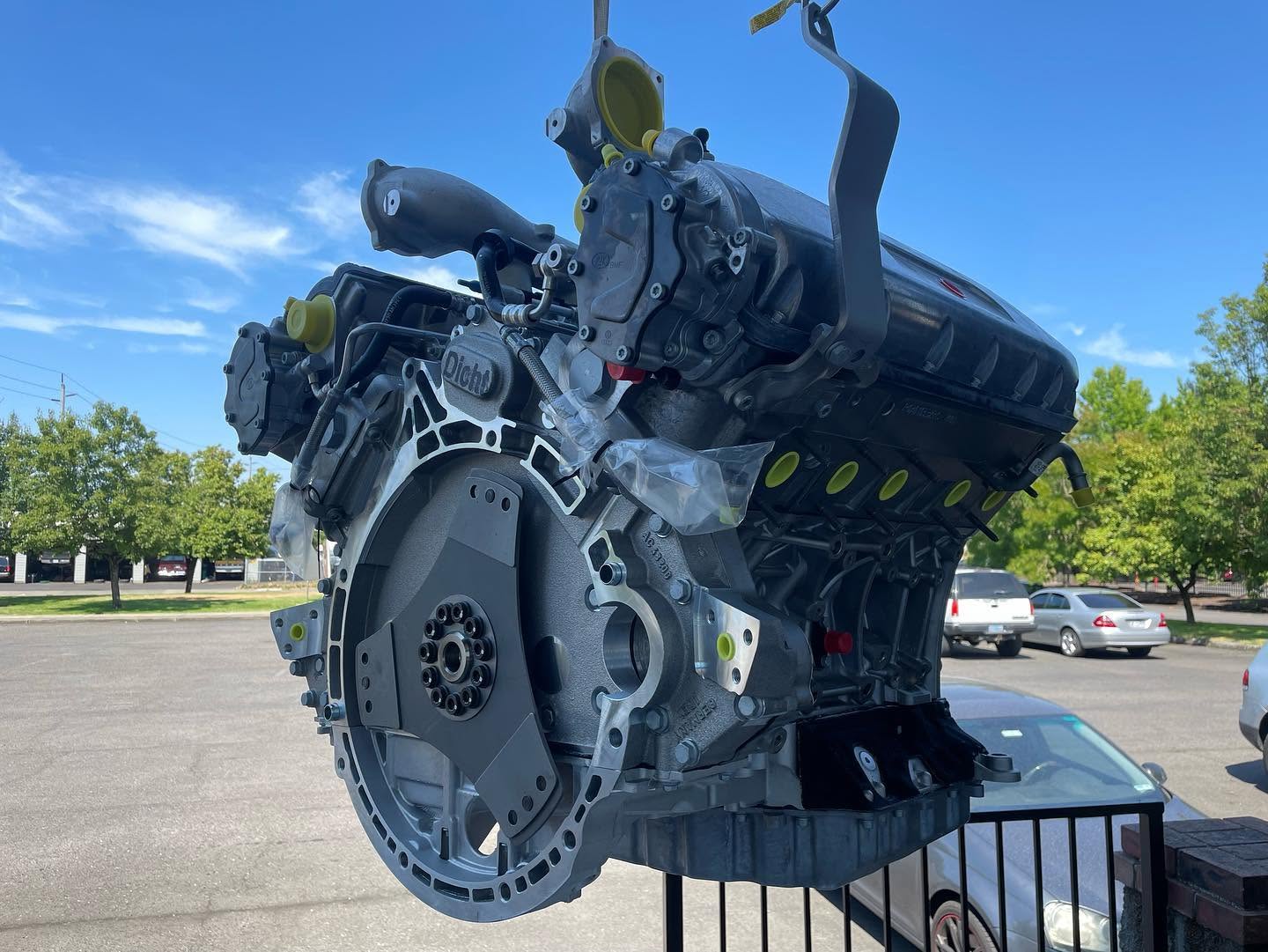 A large car engine is placed outdoors on a black metal stand, with trees and parked cars in the background.