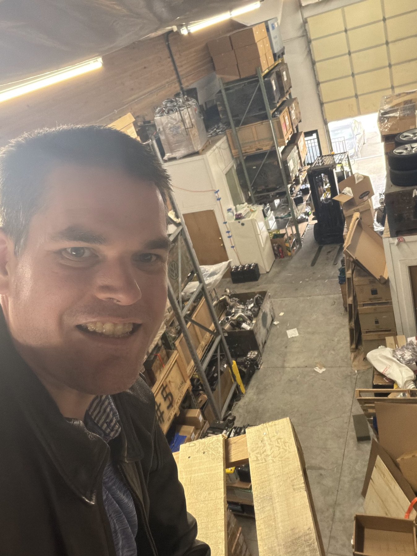 Person smiling in a warehouse with shelves and boxes, looking at the camera, with warehouse equipment in the background.