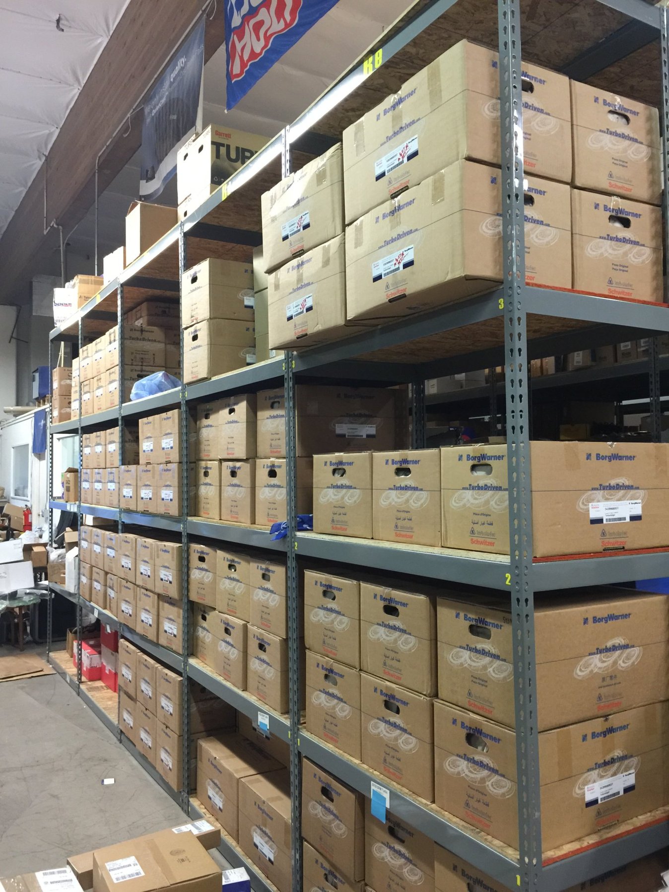 Shelves filled with stacked cardboard boxes in a warehouse, organized on metal racks, with some labels visible.