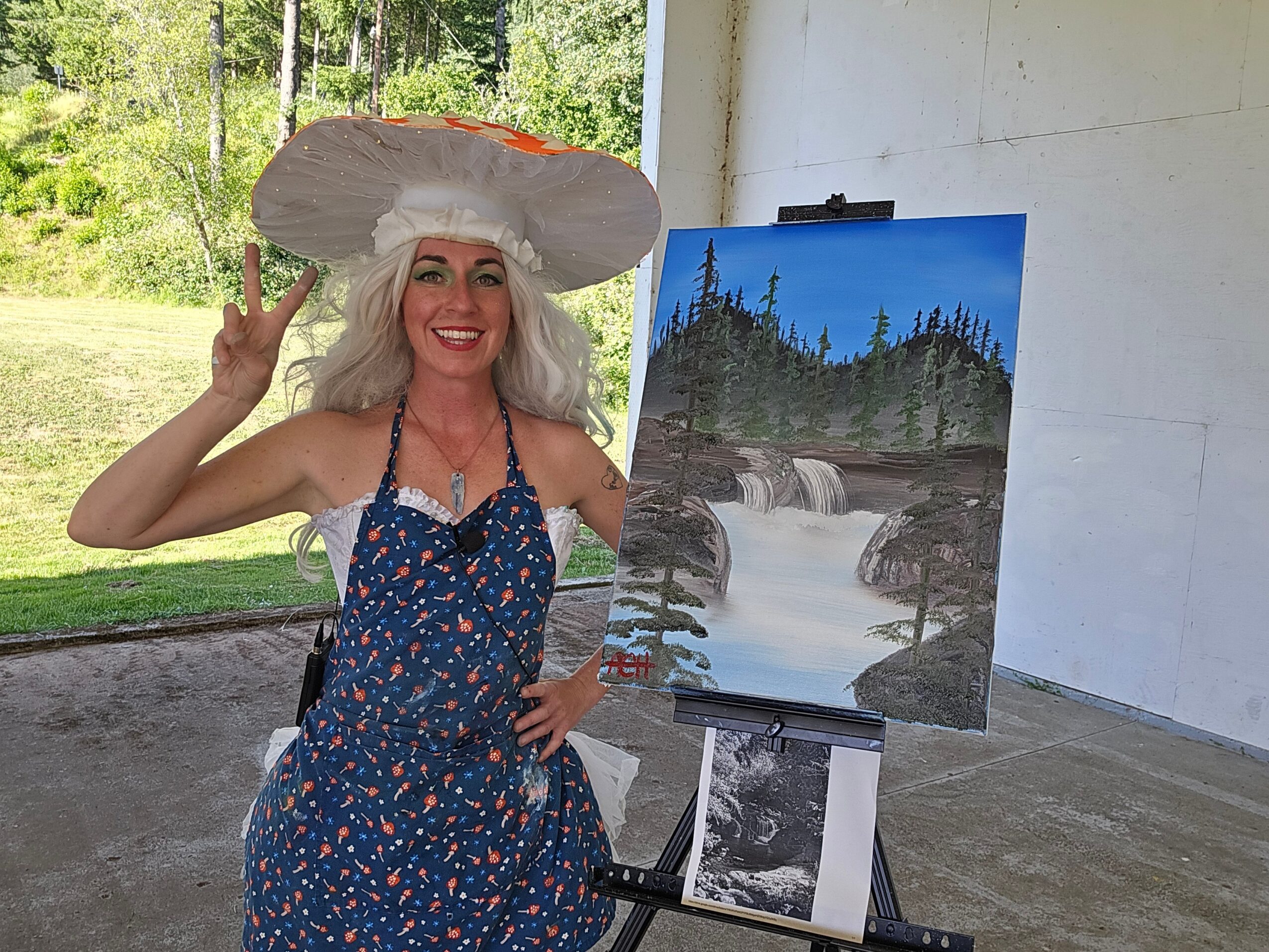 Woman with long blonde hair wearing a large hat and a blue dress with a floral pattern, smiling outdoors next to a painting of a river and trees.