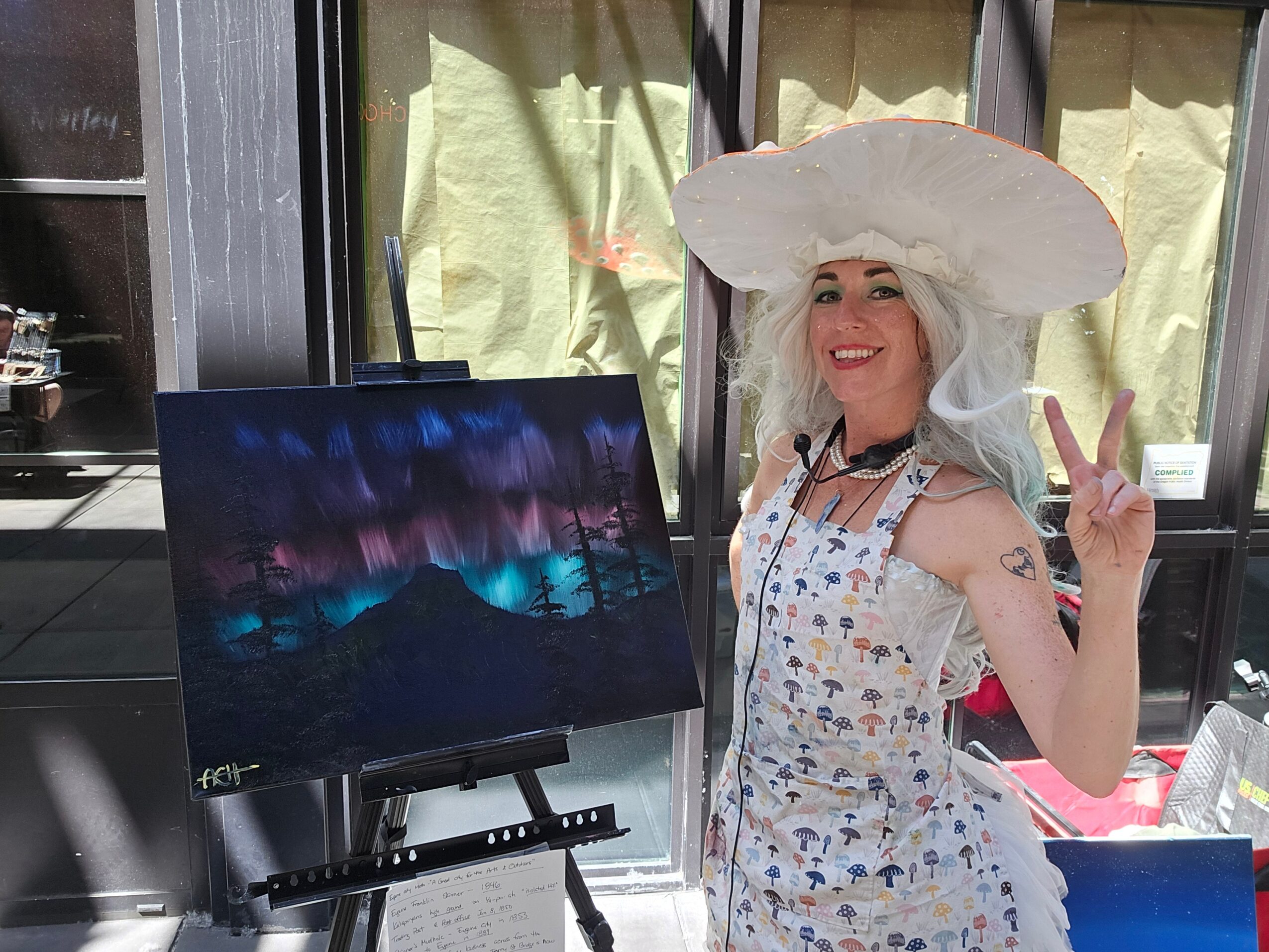 Woman with white hair and large hat making a peace sign next to a painting of a colorful night sky.