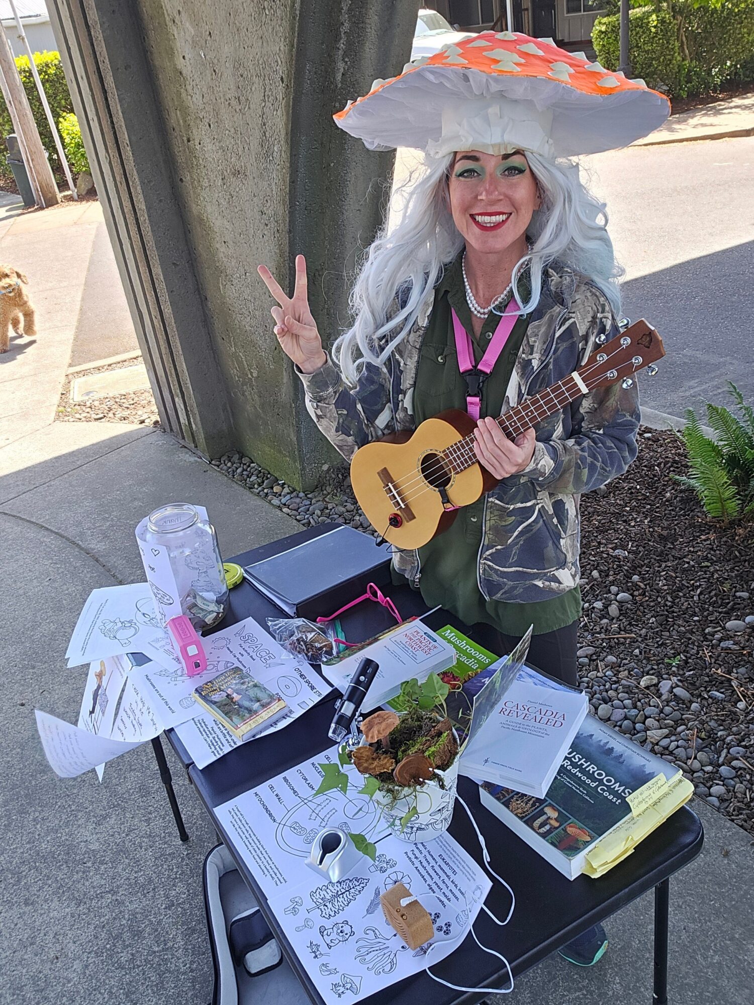 Person with long white hair wearing a large hat, playing guitar and making a peace sign outdoors.