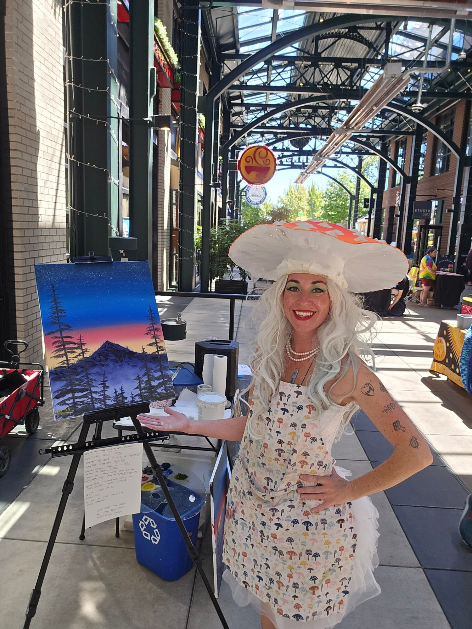 Woman with long white hair in a floral dress holding a drink, standing outdoors near a table with a colorful display.