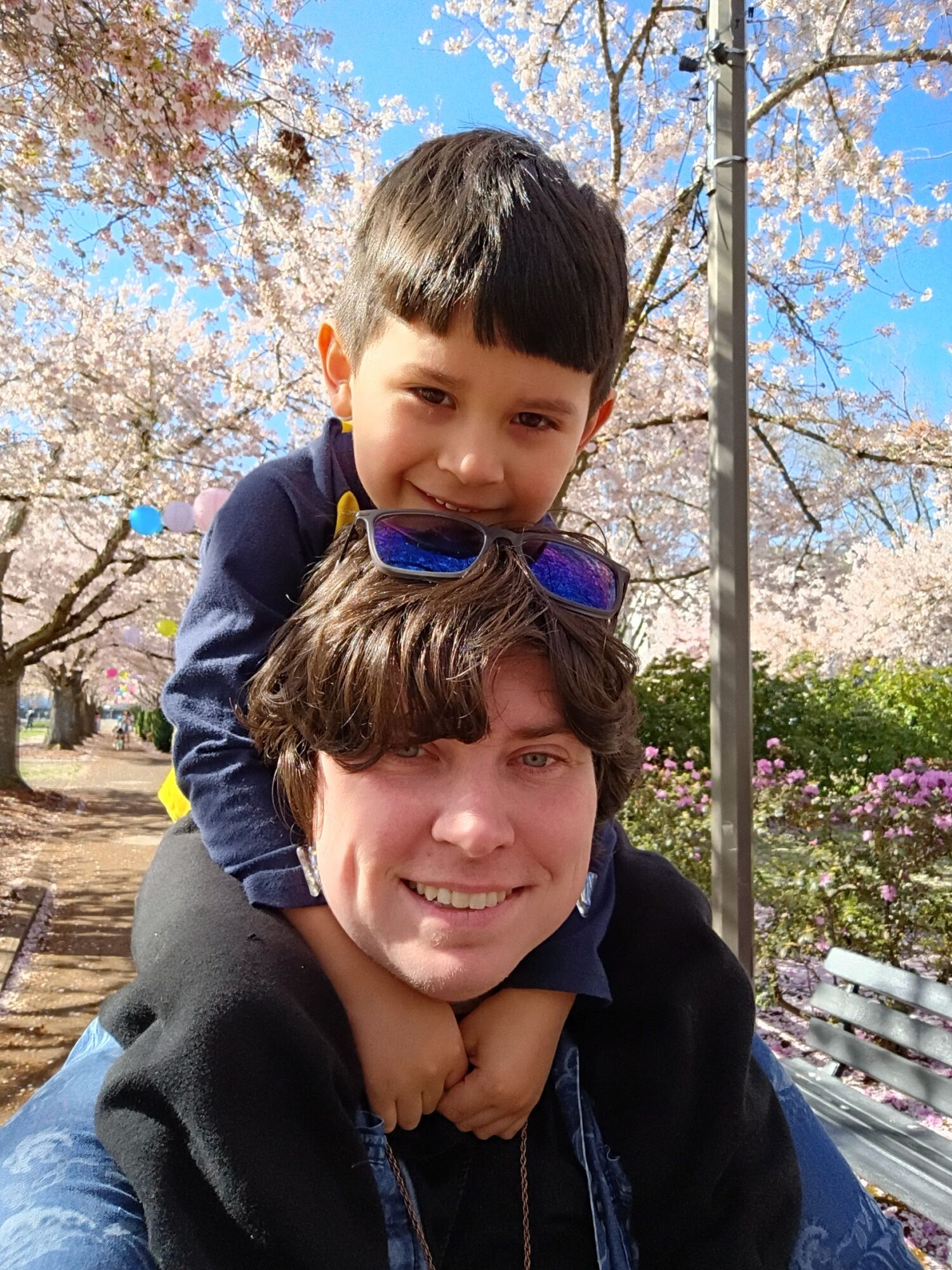 Two children smiling outdoors with blooming cherry blossoms and blue sky in background.