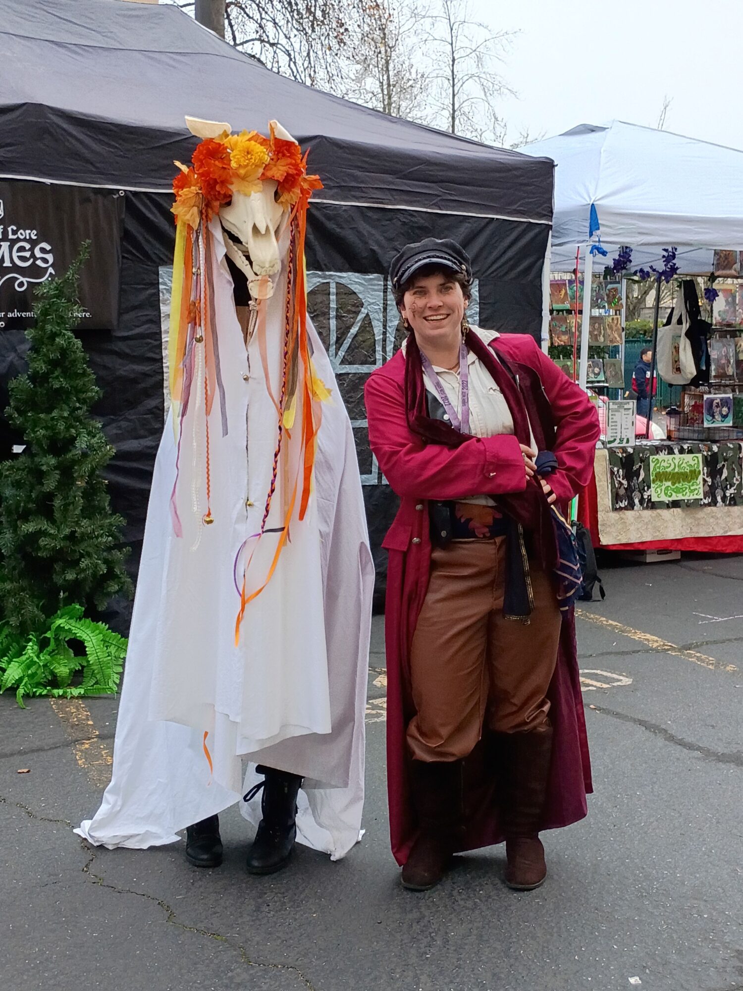 Person standing next to a decorated figure with orange and yellow ribbons, outdoors at a market or fair.