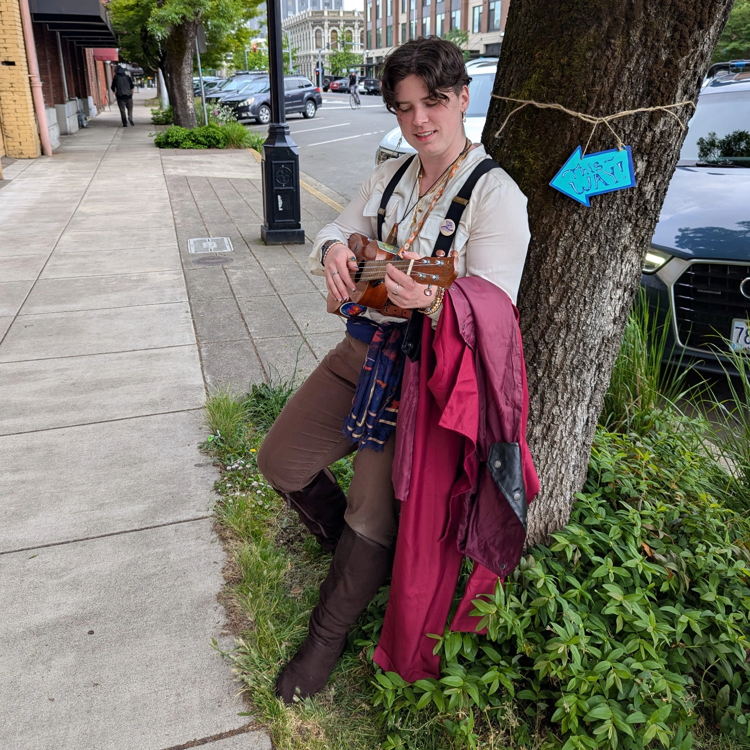 Young person leaning against a tree on a city sidewalk, holding a guitar and wearing a pink jacket. Blue arrow points to the tree.