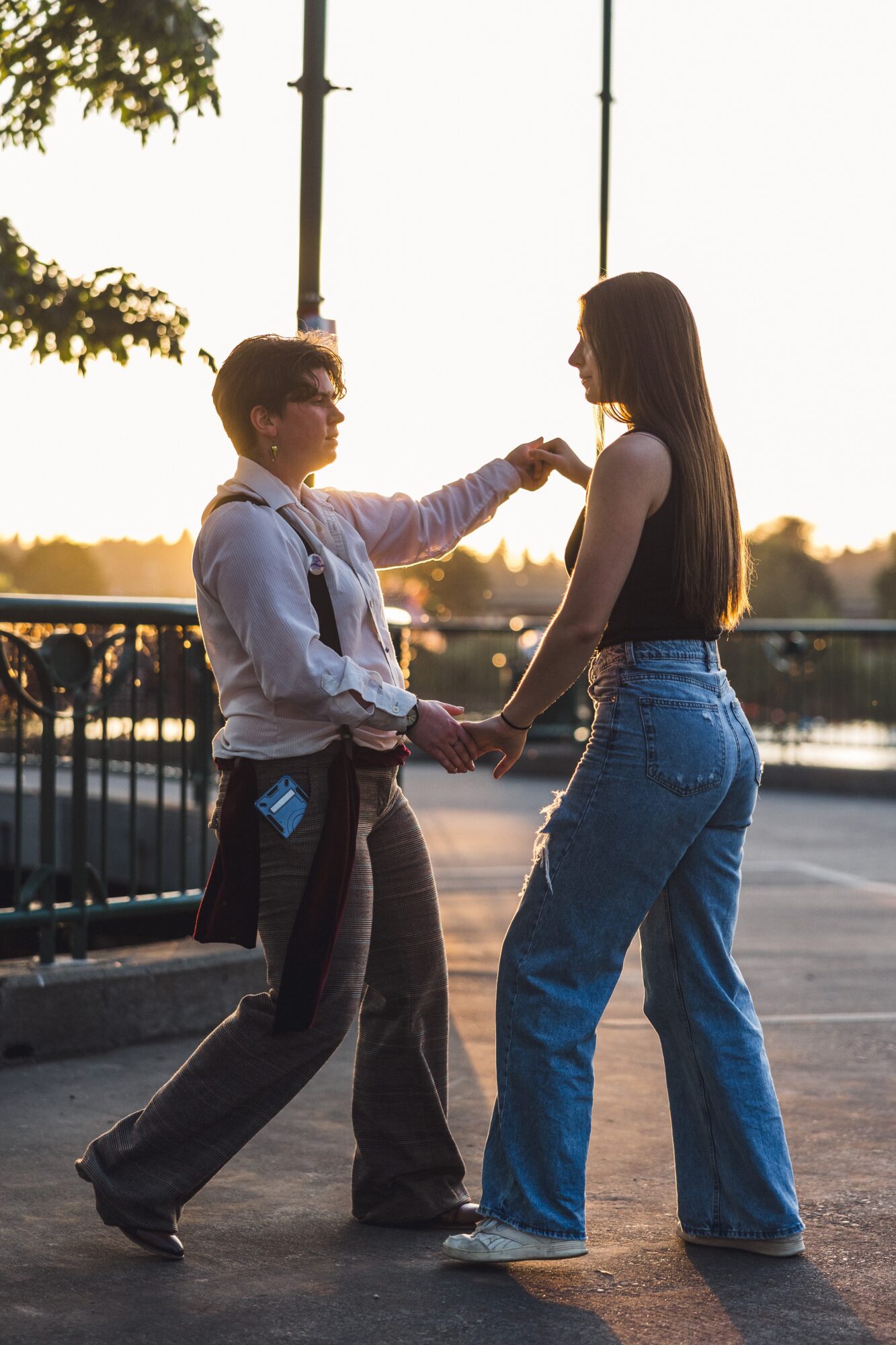 Two people holding hands and facing each other outdoors during sunset, with a railing and trees in the background.