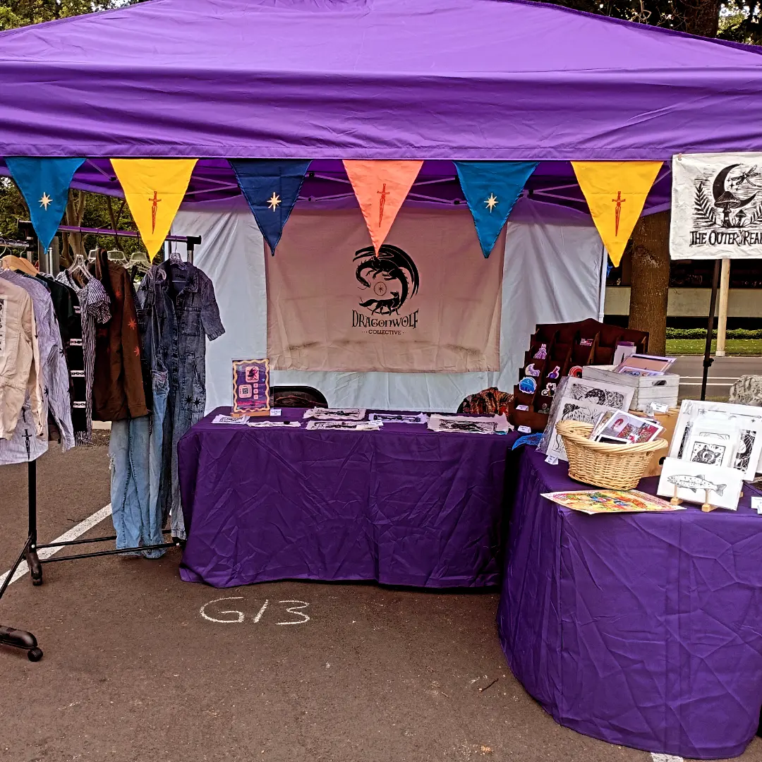 Outdoor market stall with purple canopy, displaying clothing and books, decorated with colorful triangular flags, and a table covered with purple cloth.