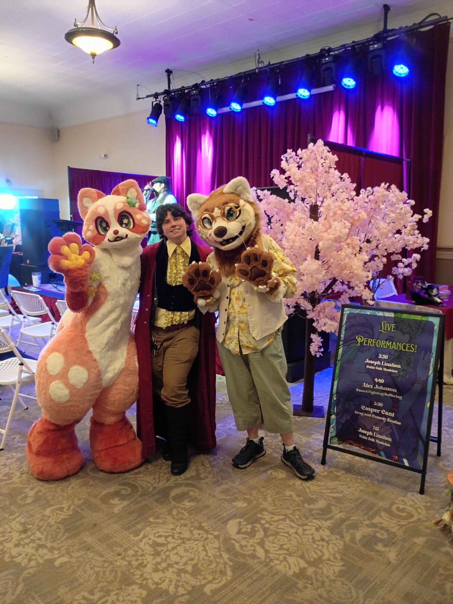 Two people in animal costumes and a woman stand in front of a stage with pink cherry blossom tree and purple curtains.