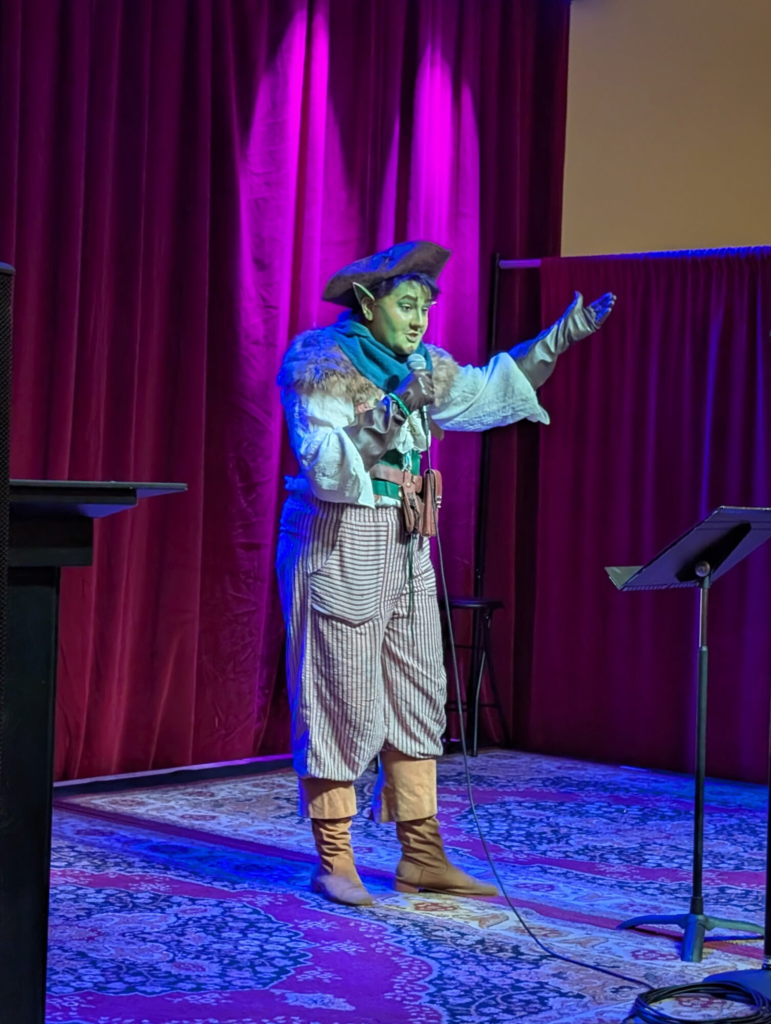 Performer dressed as a clown or mime on stage with red curtains, gesturing with one hand, standing on a patterned rug.