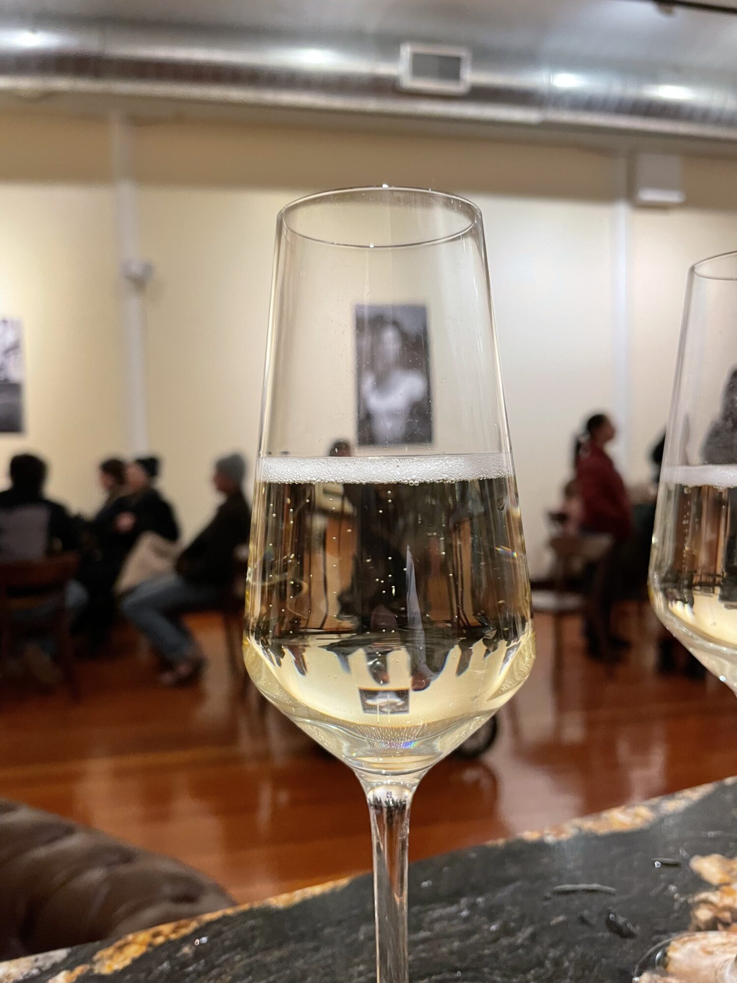 A glass of sparkling wine or champagne on a table with a blurred background of people sitting indoors.
