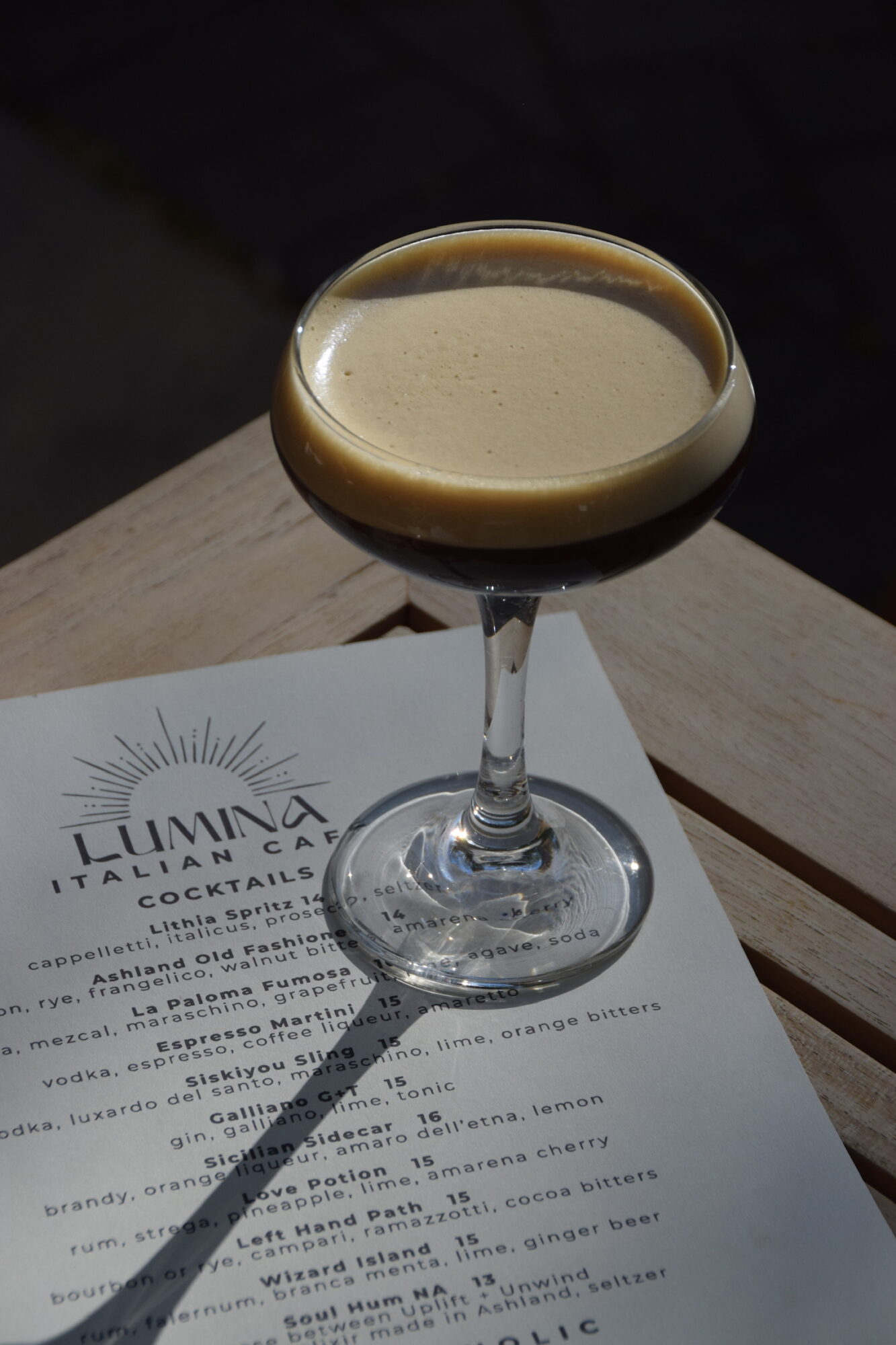 A cocktail in a glass with a dark liquid and foam on top, placed on a menu on a wooden surface.