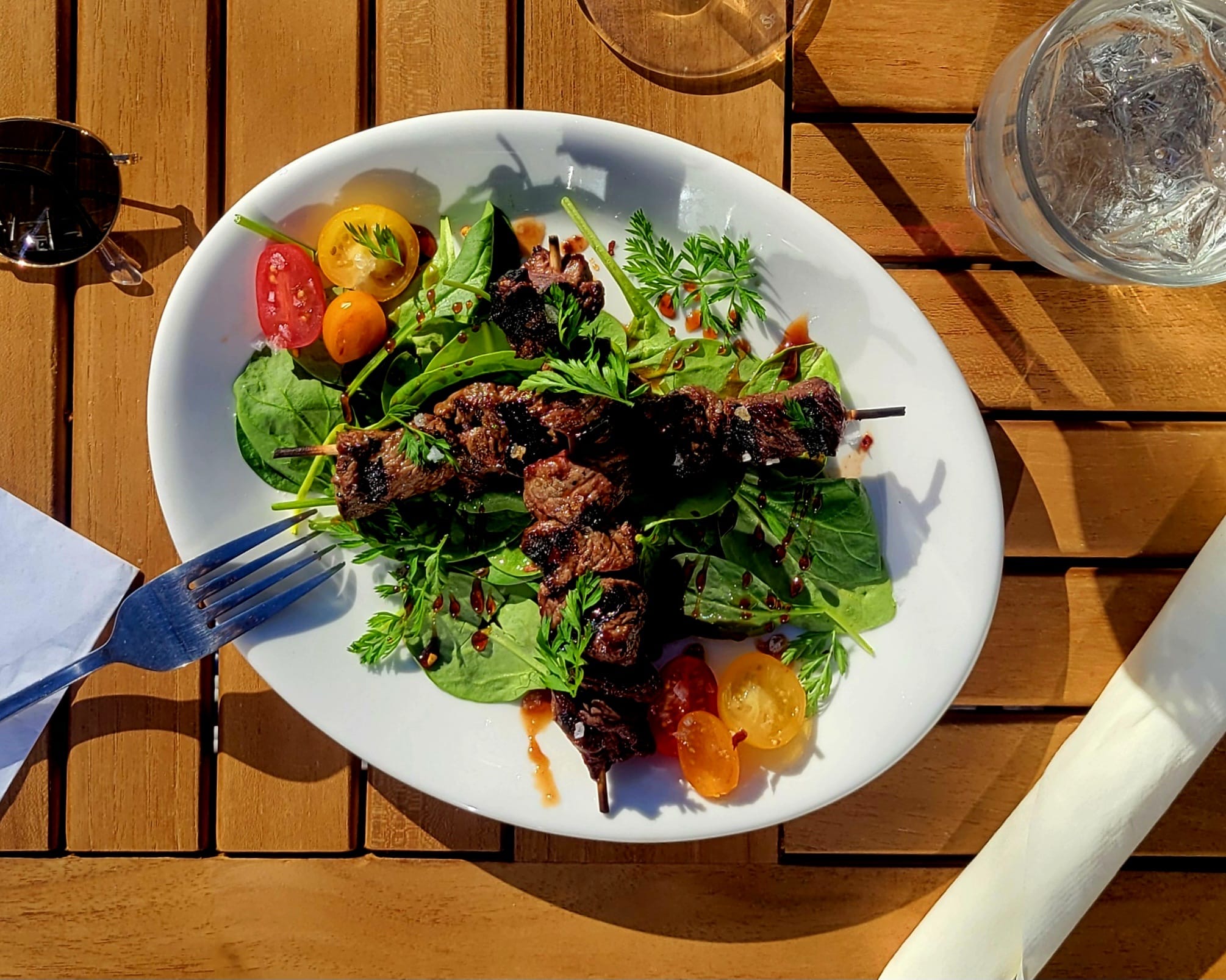 Plate with grilled meat and mixed greens, cherry tomatoes, and yellow vegetables on a wooden table, with a fork on the left.