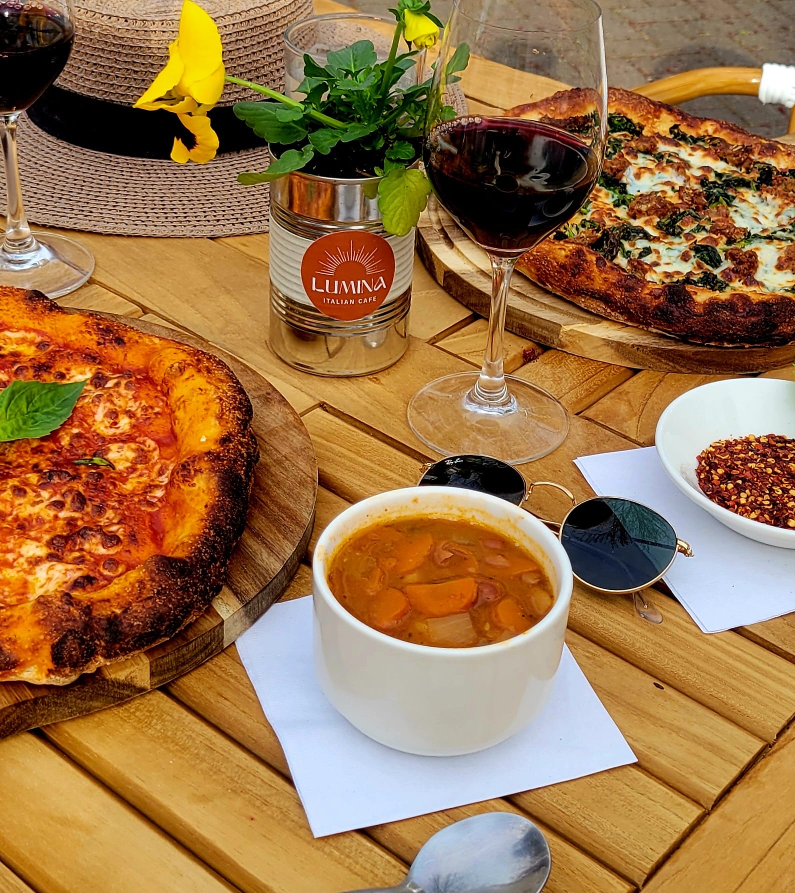 Two pizzas on wooden boards, a glass of red wine, a cup of soup, and condiments on a wooden table.