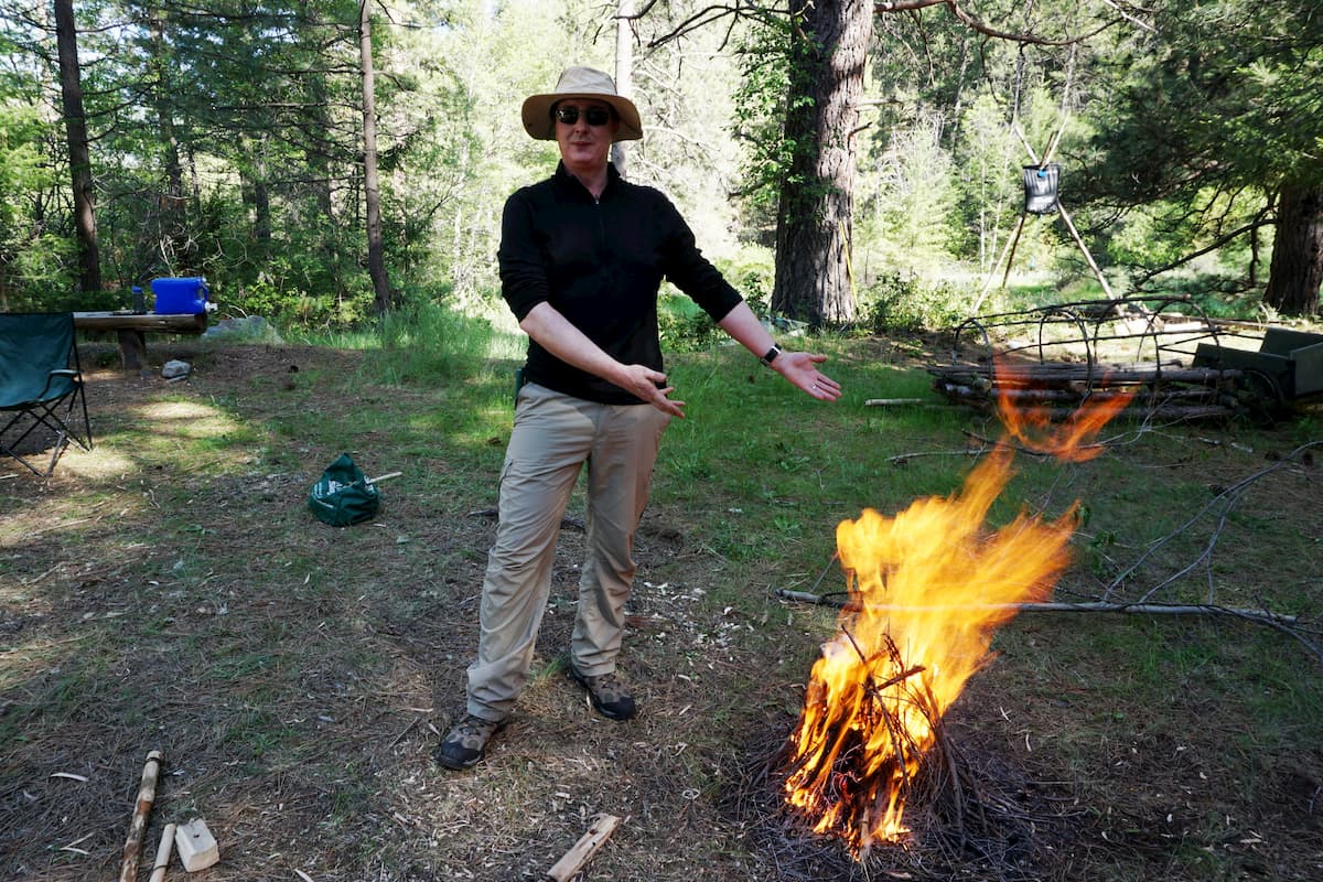 Person wearing sunglasses, hat, black shirt, and beige pants standing outdoors near a fire with trees in background.