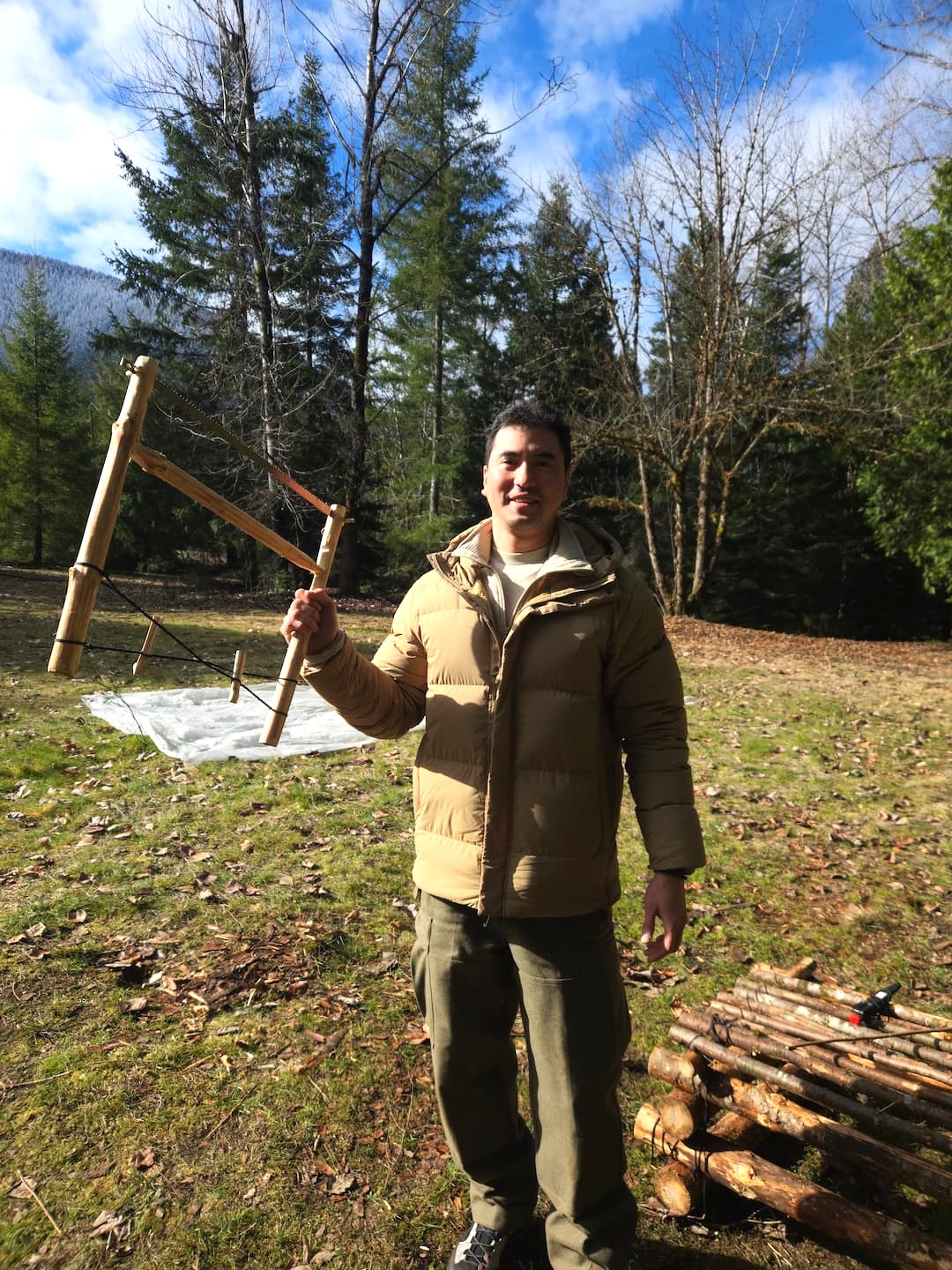 Person standing outdoors near a wooden well with a pulley system, surrounded by trees and grass, under a partly cloudy sky.