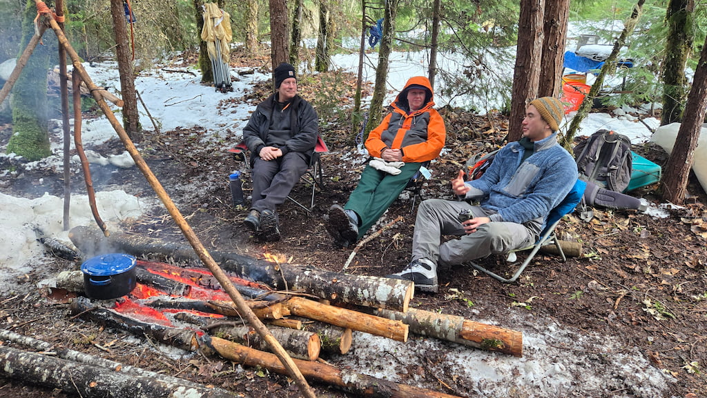 Three people sitting around a campfire in a forest, with trees and camping gear nearby.