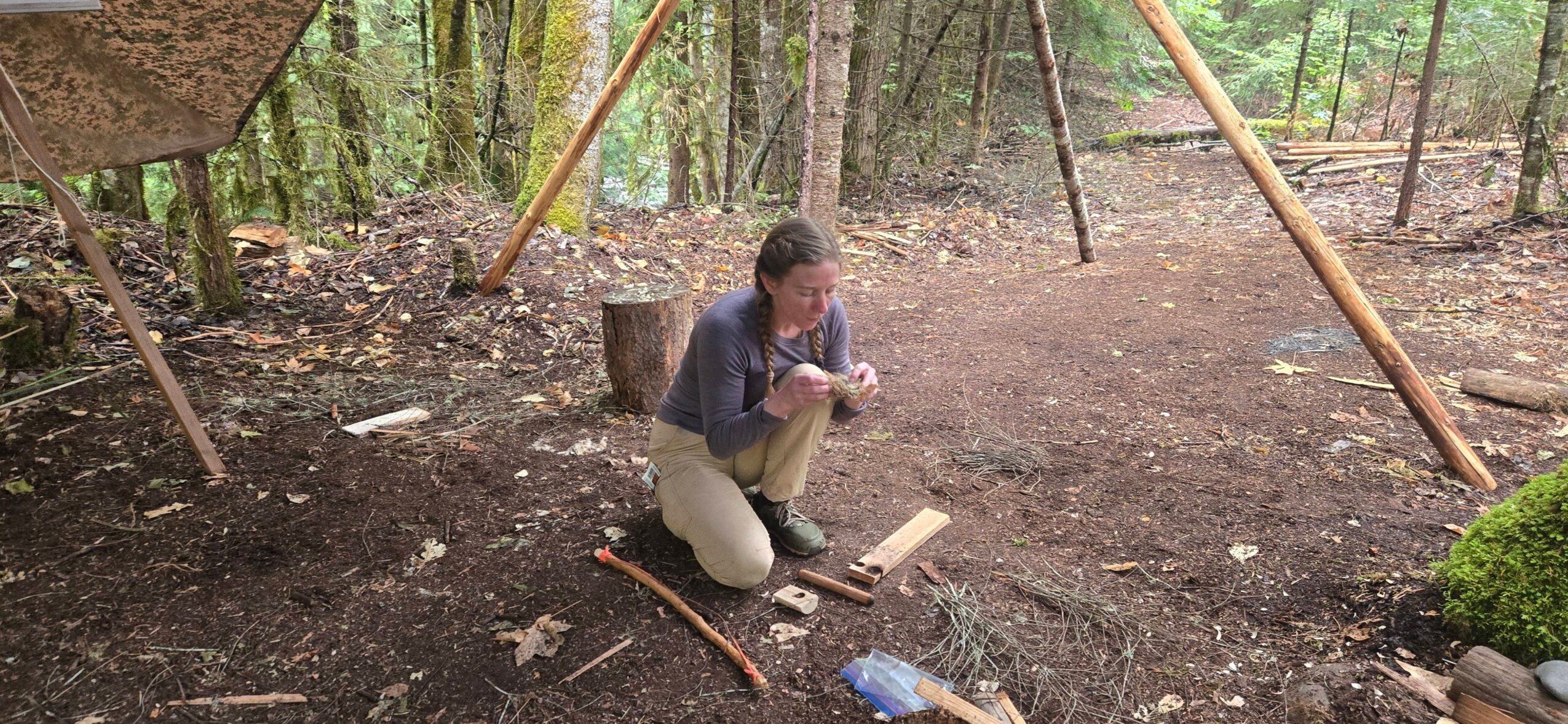 Person crouching on ground in a forested area with trees and wooden structures nearby.