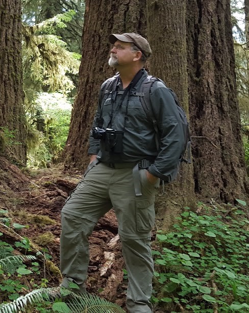 Man in outdoor clothing standing in a forest with large trees and green foliage.