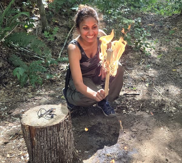 Young woman outdoors holding a large burning torch, smiling, with a tree stump nearby and greenery in the background.