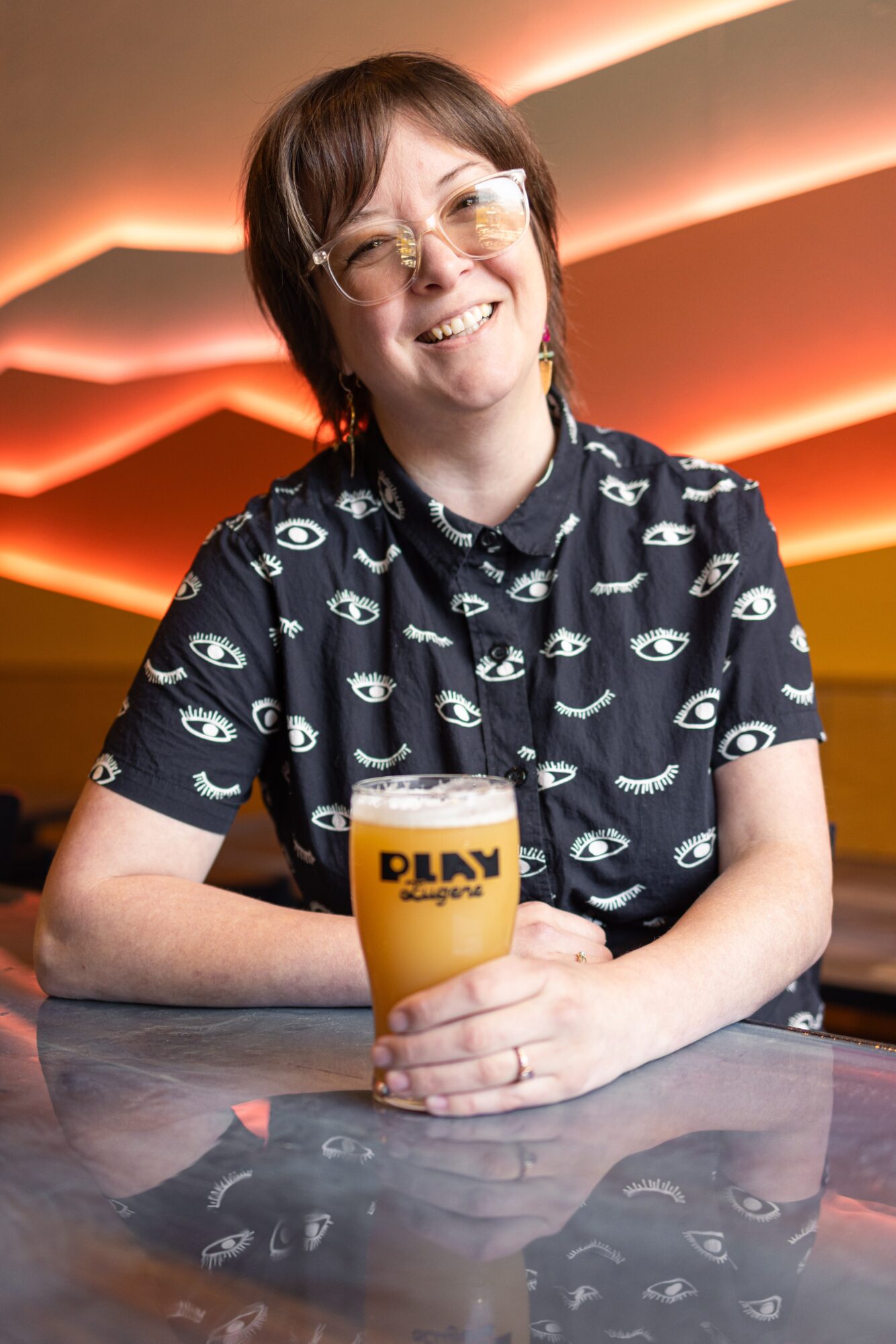 Person with short brown hair and glasses smiling, holding a yellow drink in a bar or restaurant setting.