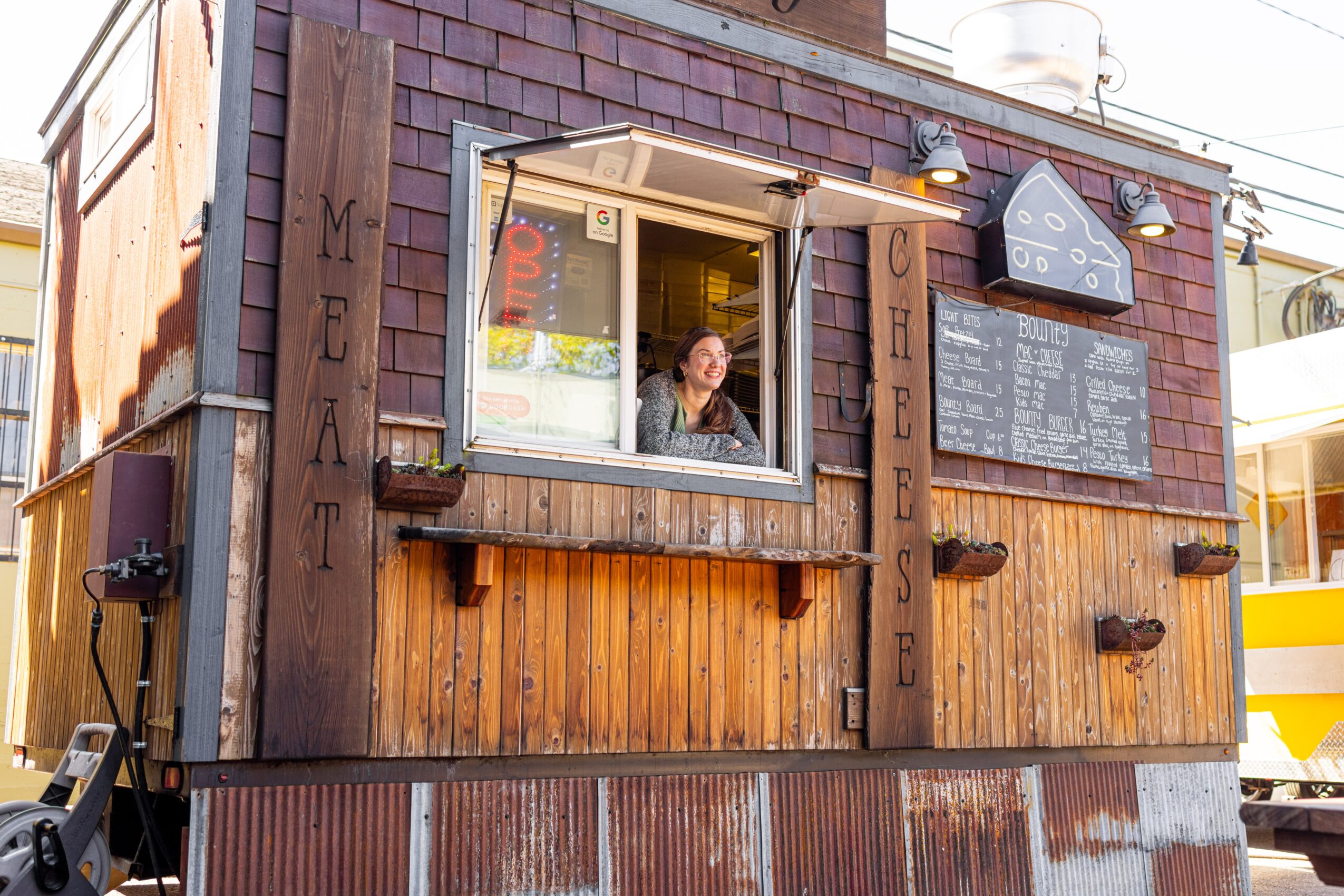 Small wooden food stand with a person inside, menu board, and decorative signs, outdoors on a sunny day.