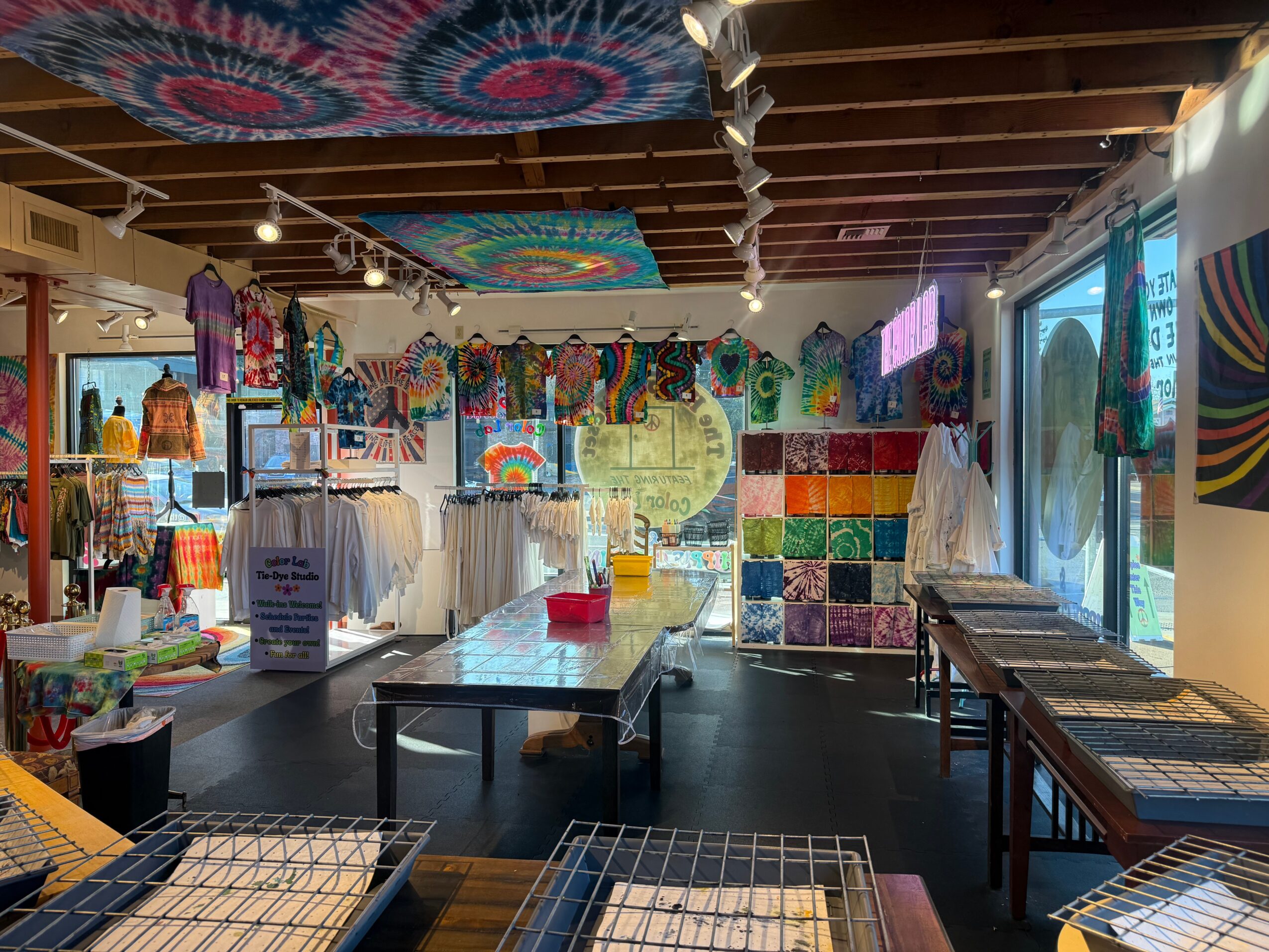 Interior of a colorful store with hanging textiles, clothing racks, tables, and large windows, decorated with vibrant tapestries and artwork.