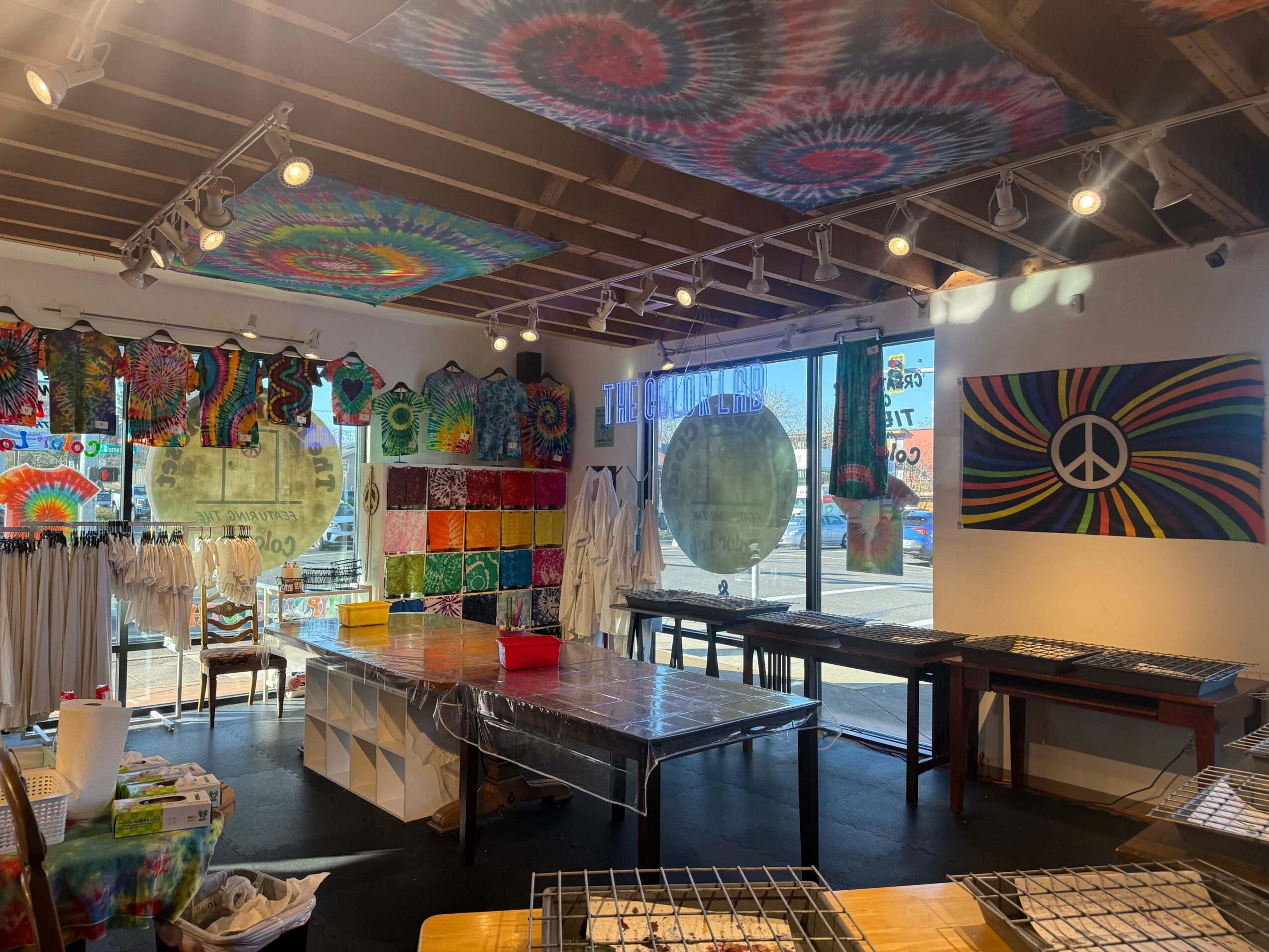 Indoor art studio with colorful paintings, fabric, and tables, large window with natural light, and ceiling decorated with tie-dye fabric.