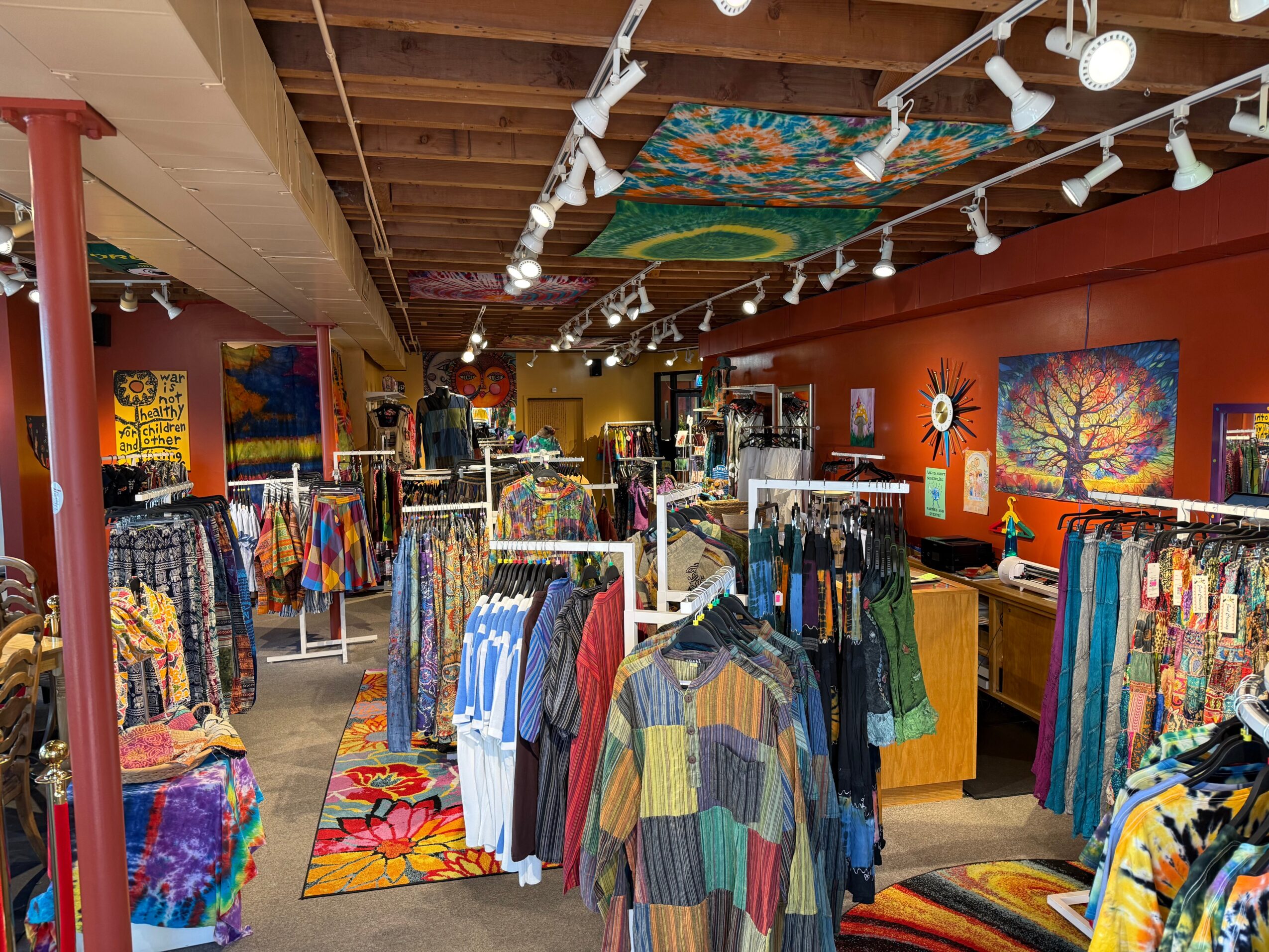 Clothing store interior with racks of colorful clothes, vibrant wall art, and ceiling artwork, illuminated by track lighting.