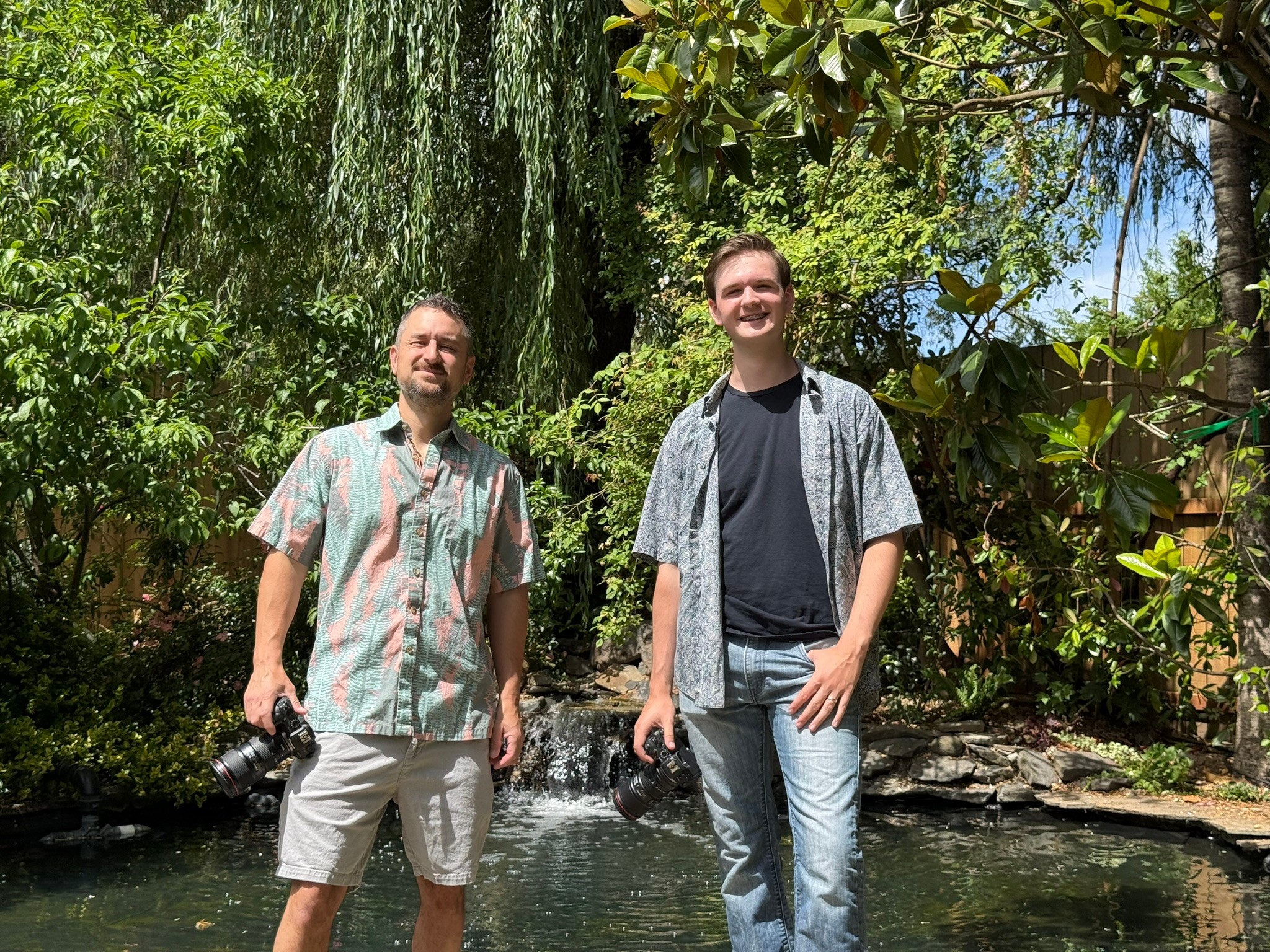 Two men stand outdoors near a pond with trees and greenery in the background, smiling at the camera.