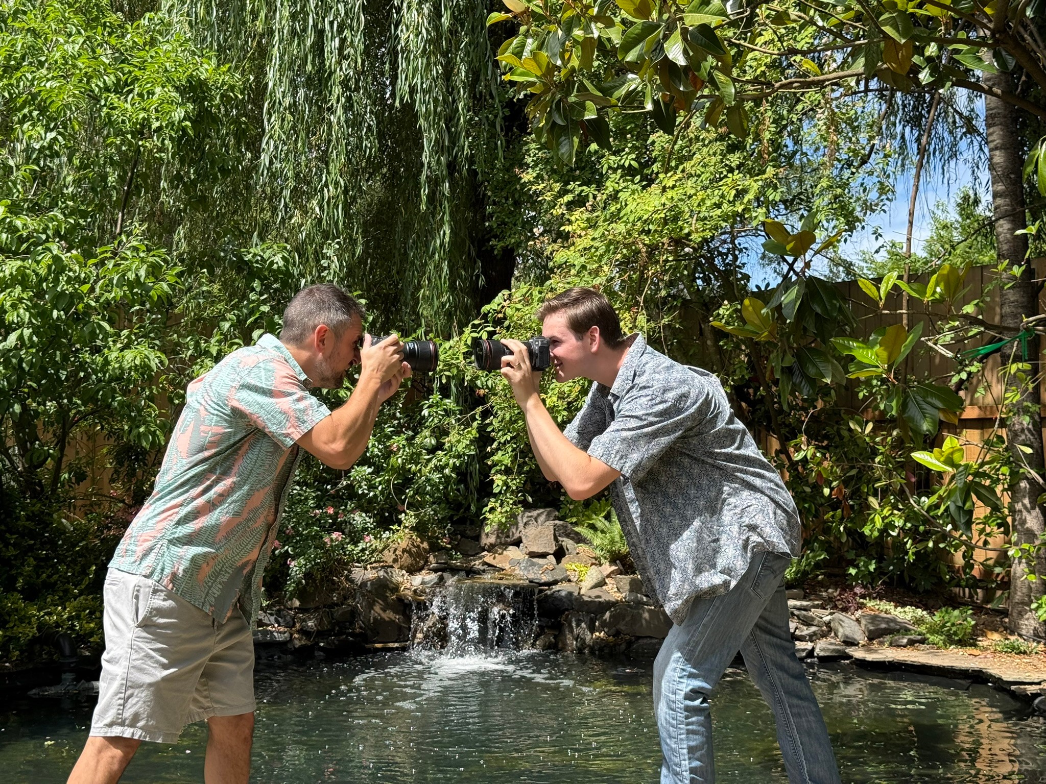 Two men taking photos of each other outdoors near a small waterfall surrounded by greenery.