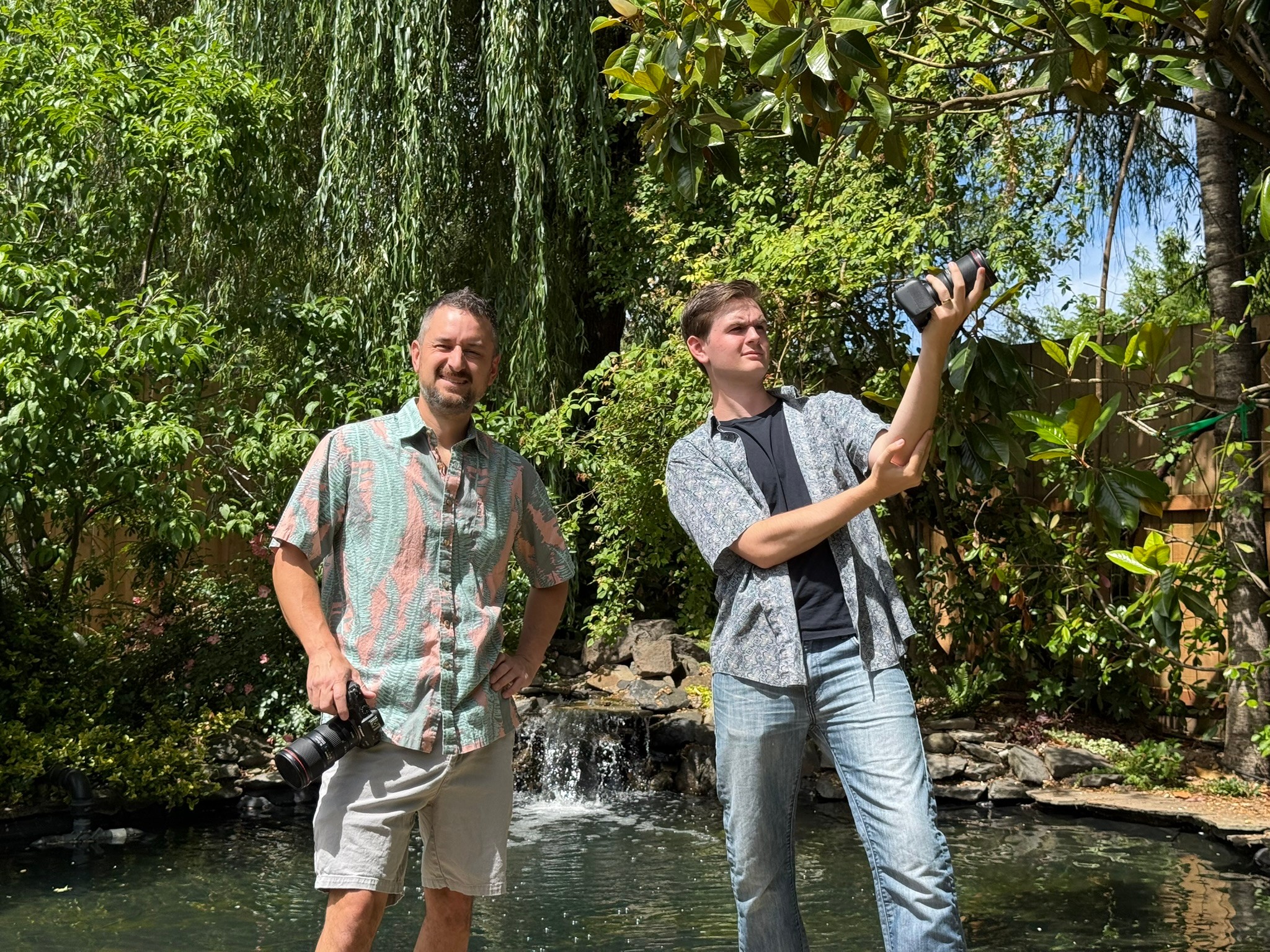 Two men standing near a small waterfall in a lush, green outdoor setting, one holding a camera and the other smiling.