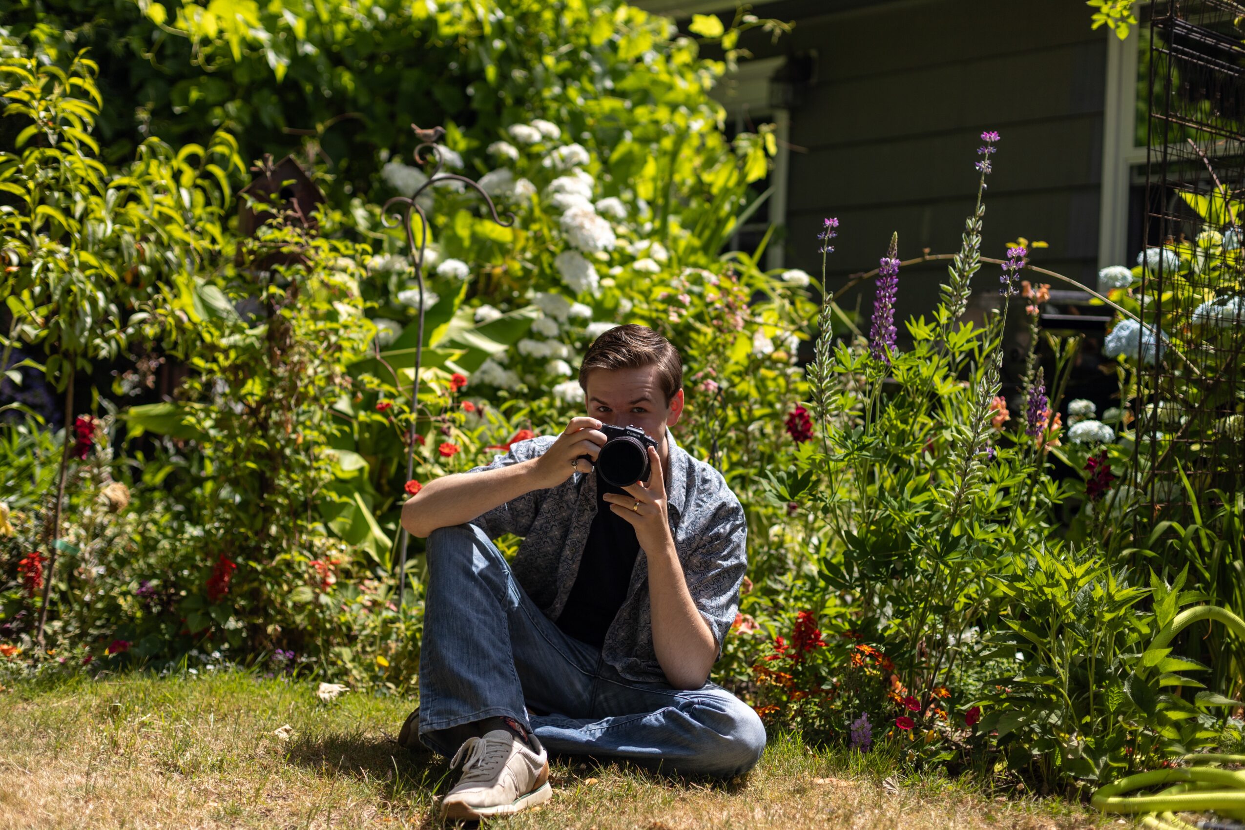 Person sitting on grass in garden, holding camera up to face, surrounded by flowers and greenery.