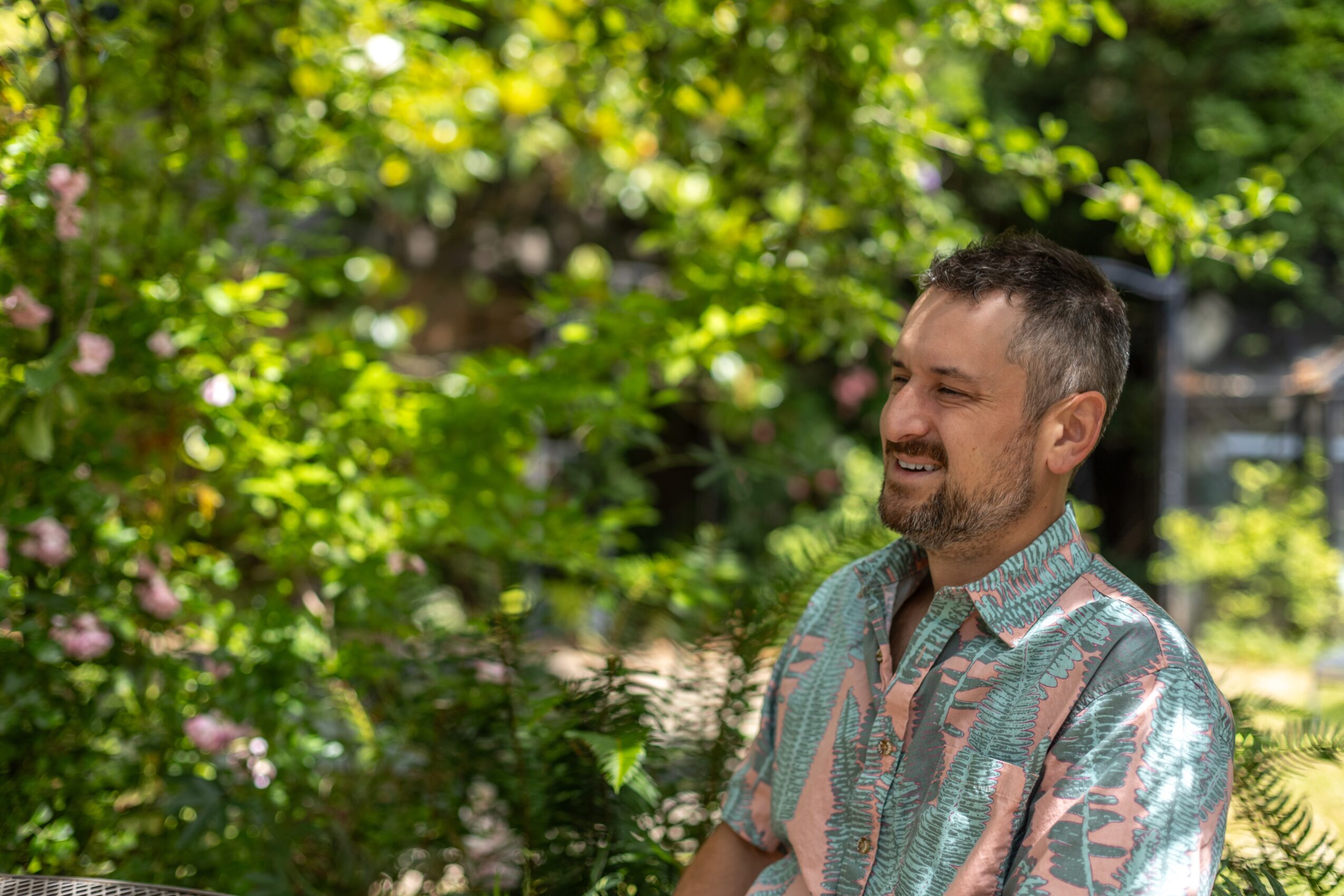 Man with short hair and beard smiling outdoors surrounded by green trees and plants.