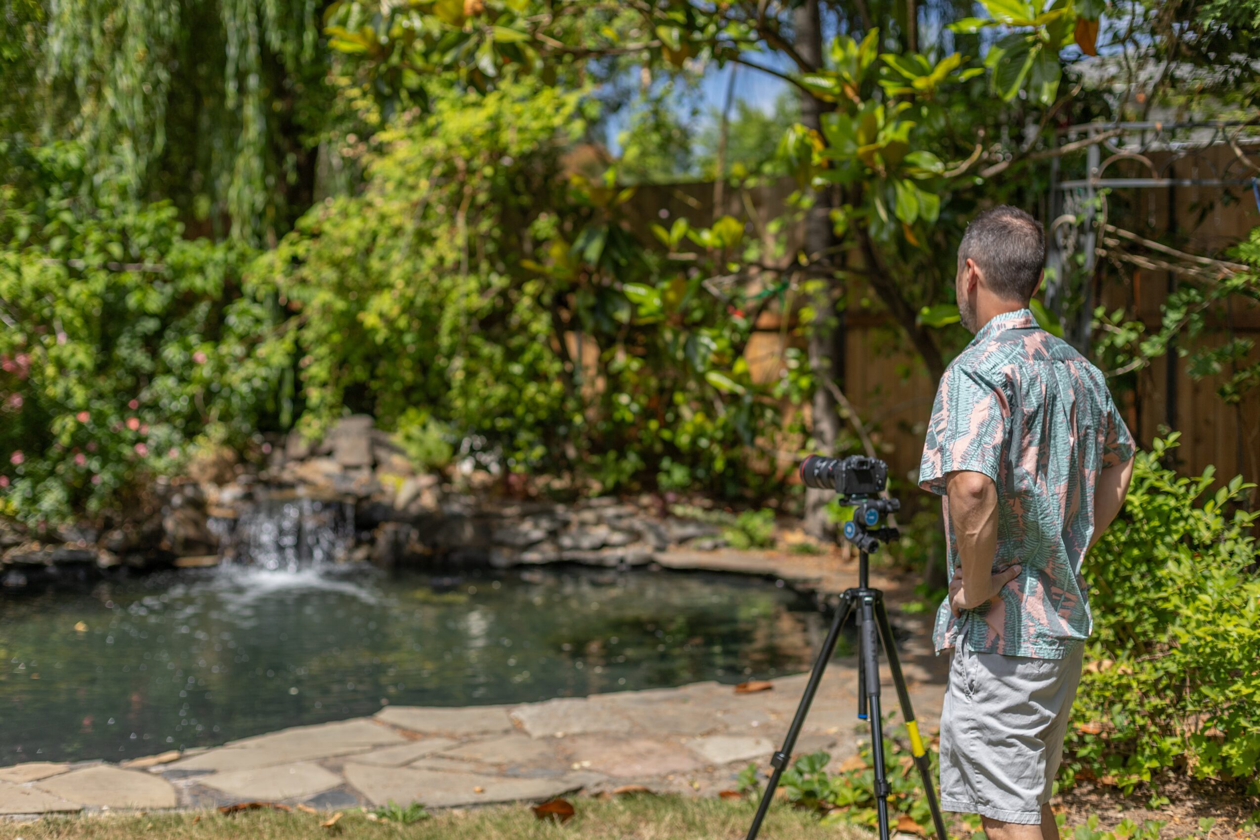 Man standing outdoors near a pond with a tripod, surrounded by trees and greenery.