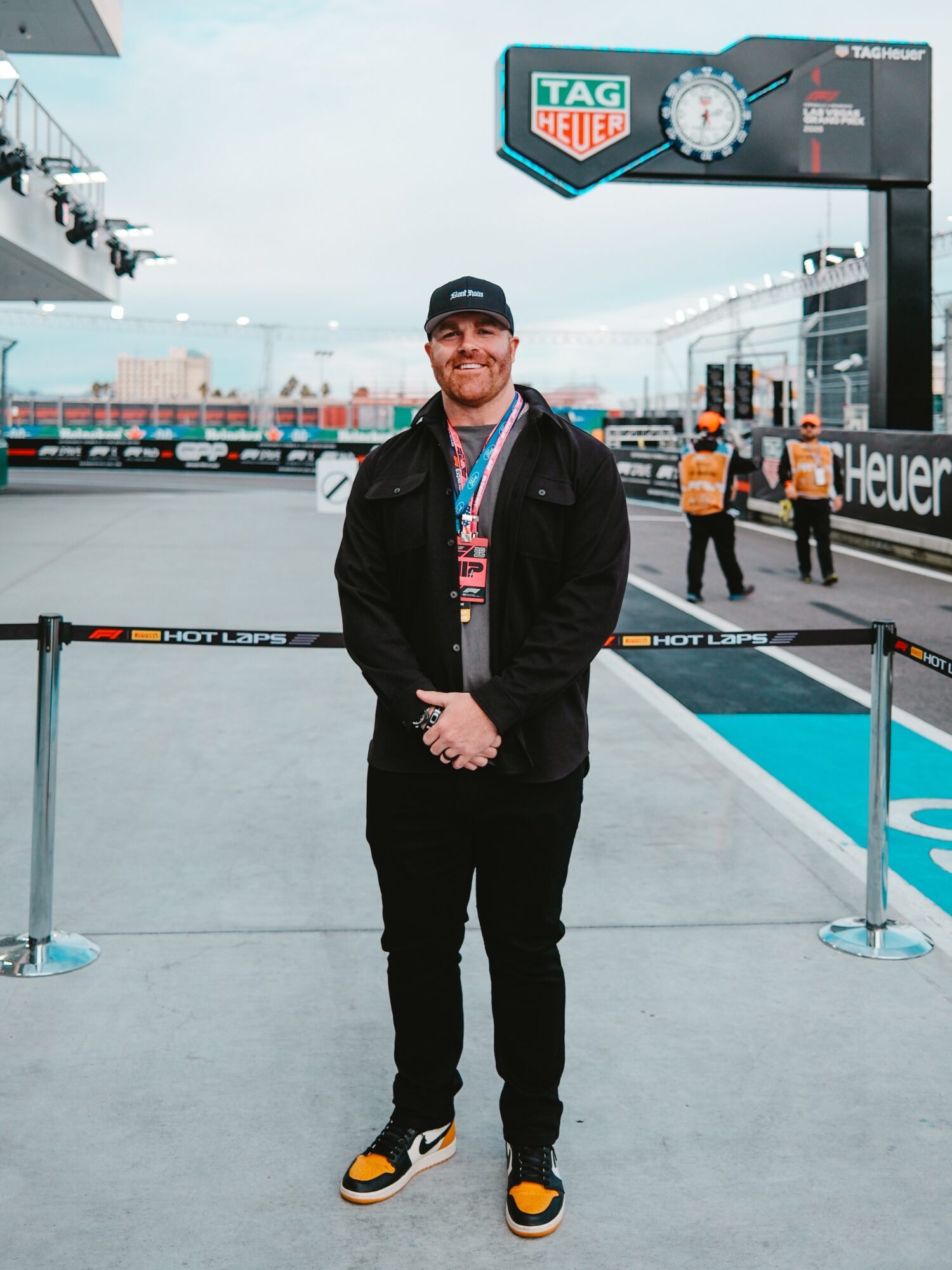 Man standing outdoors at a racing event, smiling, wearing a black jacket, black pants, and sneakers, with race track and signage in background.