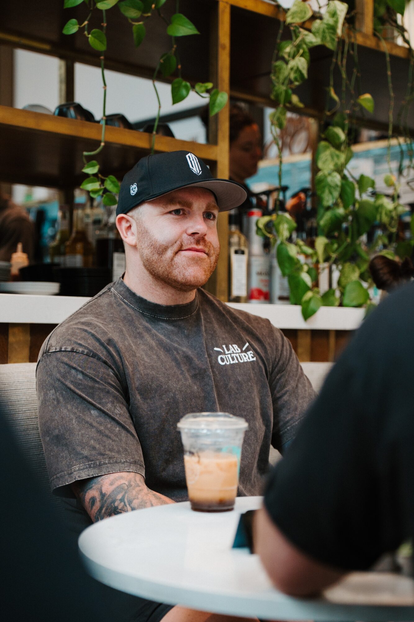 Man with a beard and tattoos wearing a black cap and t-shirt sitting at a table with a drink, in a cafe with plants.