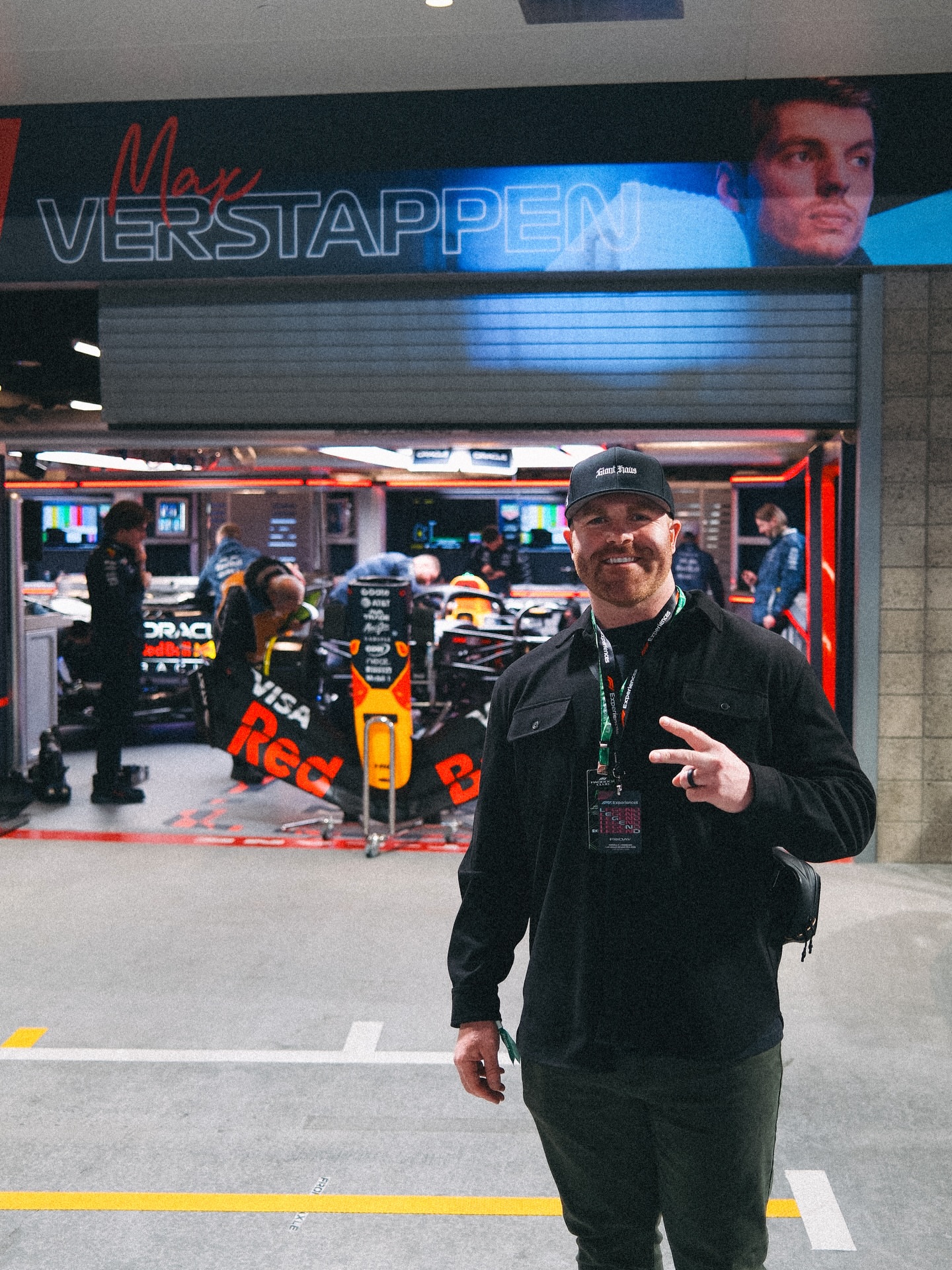 Man smiling and making a peace sign in front of a racing event with Red Bull branding, people and cars in background.