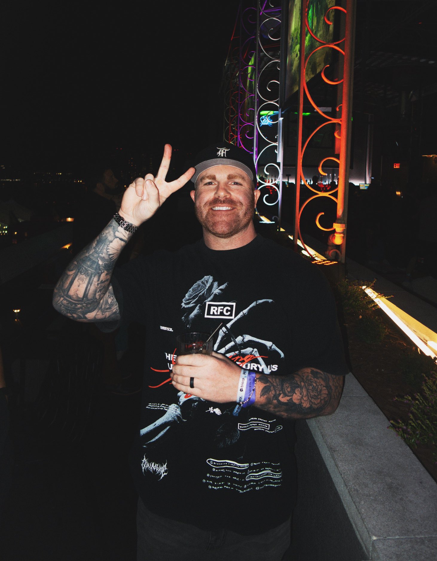 Man with tattoos smiling and making a peace sign, holding a drink, standing outdoors at night with colorful lights behind him.