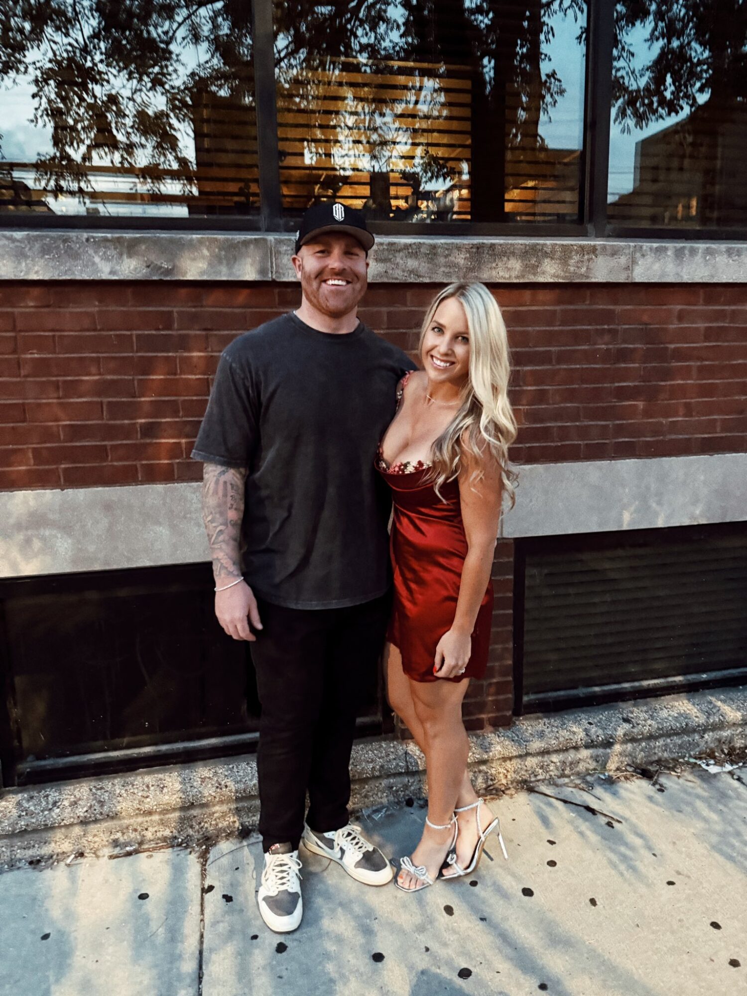 A man and woman stand together outdoors in front of a brick wall and window, smiling at the camera.