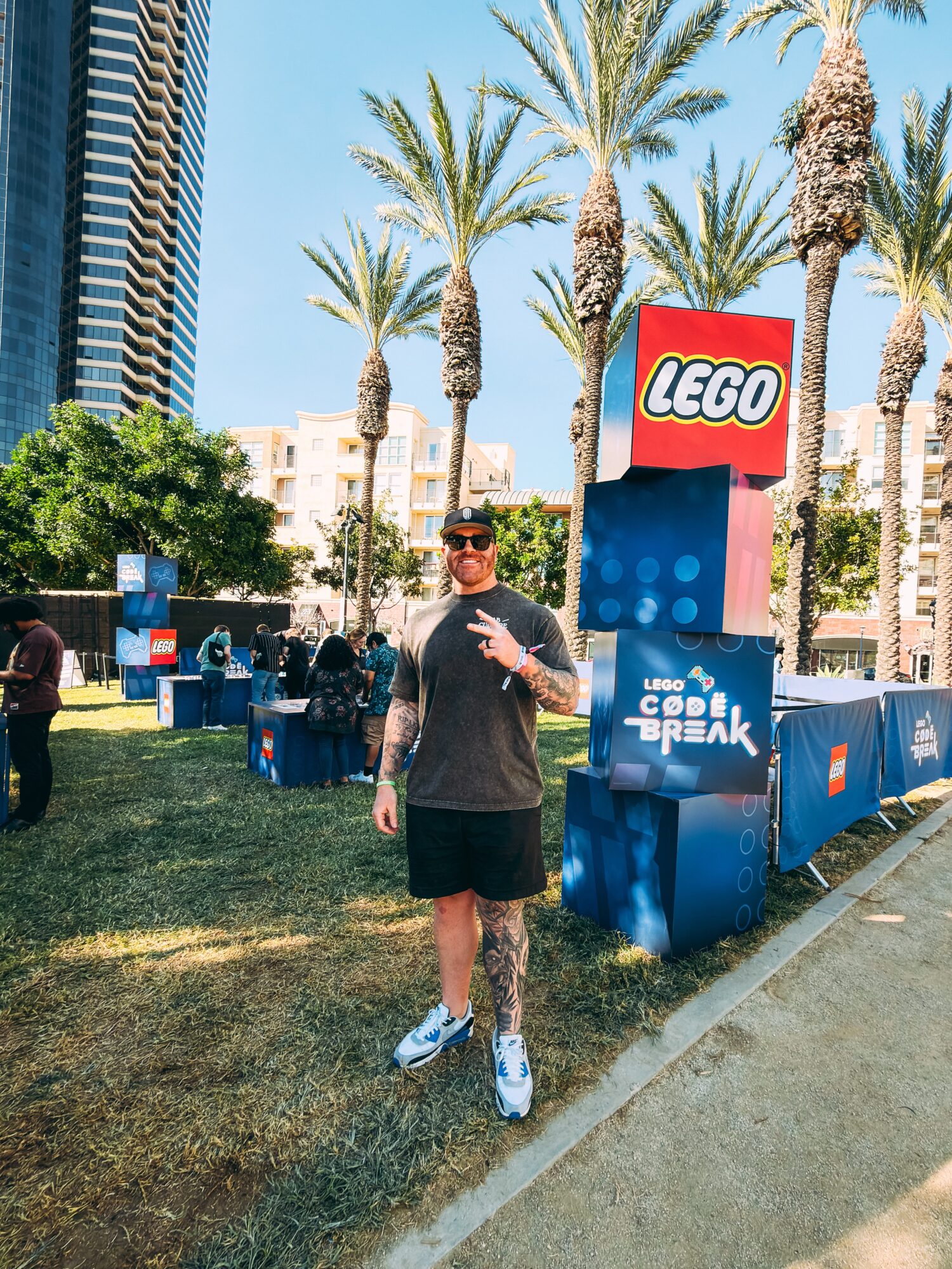 Man standing outdoors near palm trees and LEGO signage, holding a drink, wearing sunglasses, black t-shirt, shorts, and sneakers.
