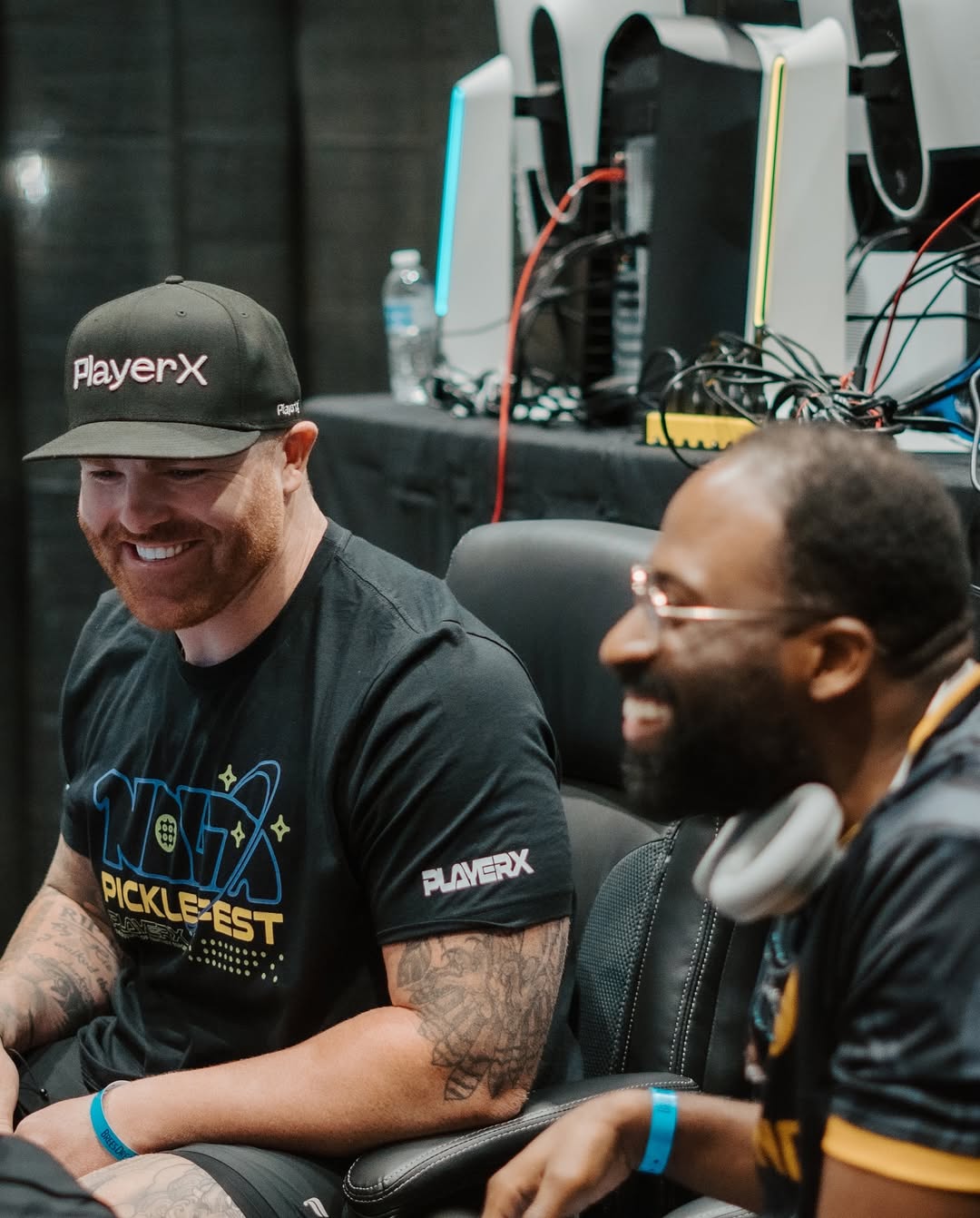 Two smiling men sitting at a gaming event with computers and monitors in the background.