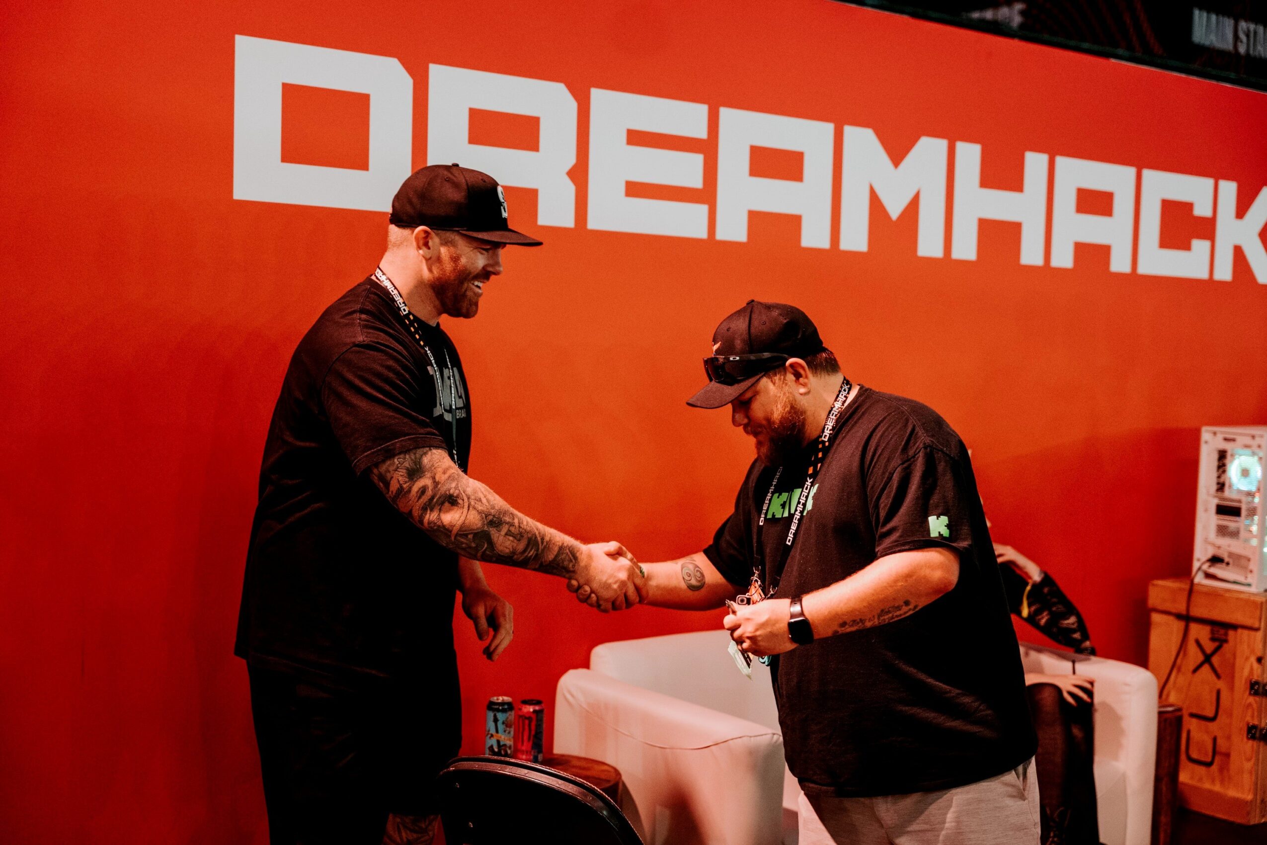 Two people shaking hands in front of a red backdrop with 'DREAMHACK' text, one with tattoos and a cap, the other with glasses.