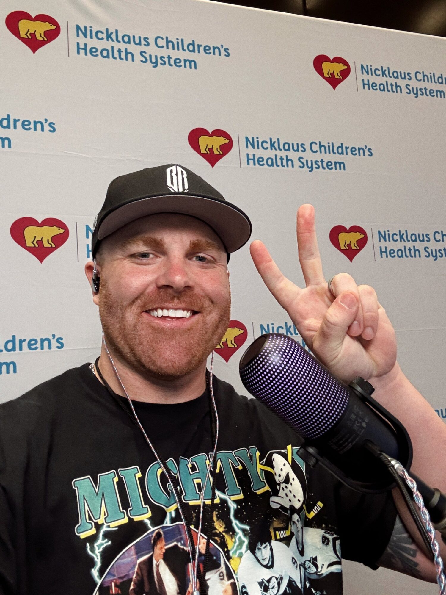 Smiling man wearing a black cap and T-shirt, holding up one finger, in front of a Nicklaus Children's Health System backdrop.