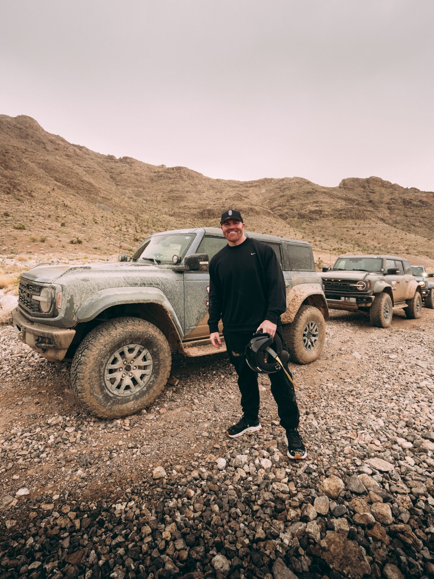 Person standing next to a rugged off-road vehicle in a desert landscape with hills in the background.