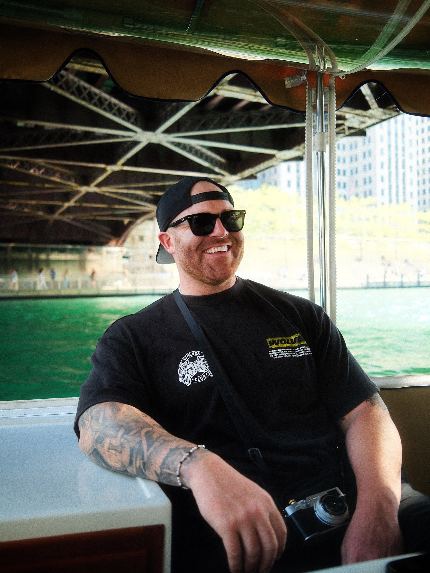 Man with sunglasses and a beard, smiling, sitting outdoors under a bridge with water and city buildings in background.