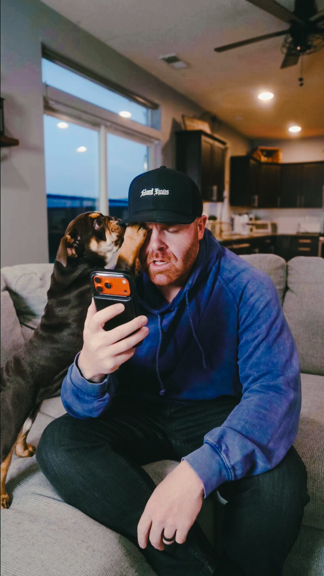 Man in blue hoodie and black cap sitting on couch with dog licking his face, holding phone, in living room.